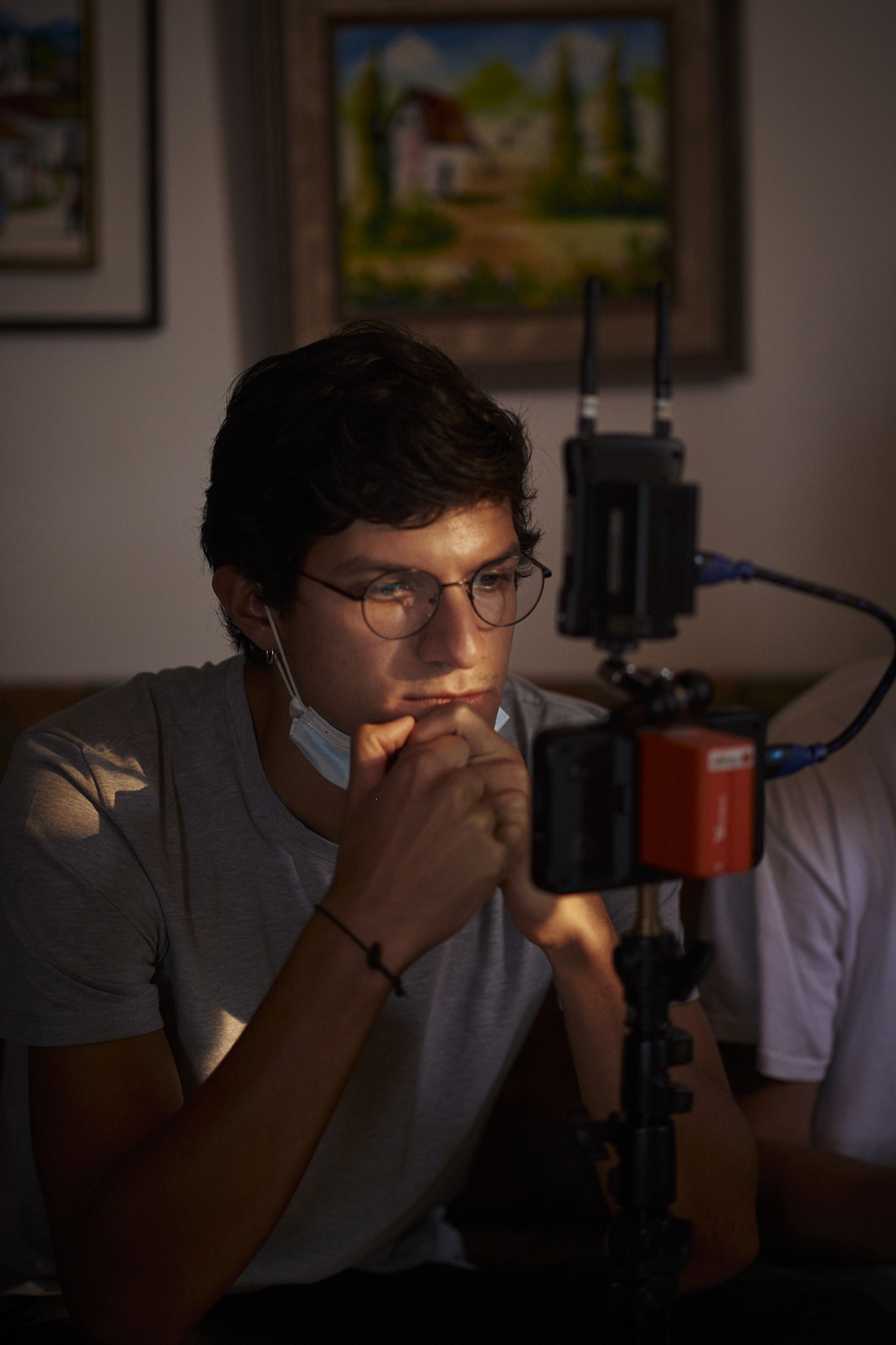 Un joven pensativo con gafas revisa el metraje en un monitor de cámara en una habitación con poca luz, con una máscara colgando de una oreja.