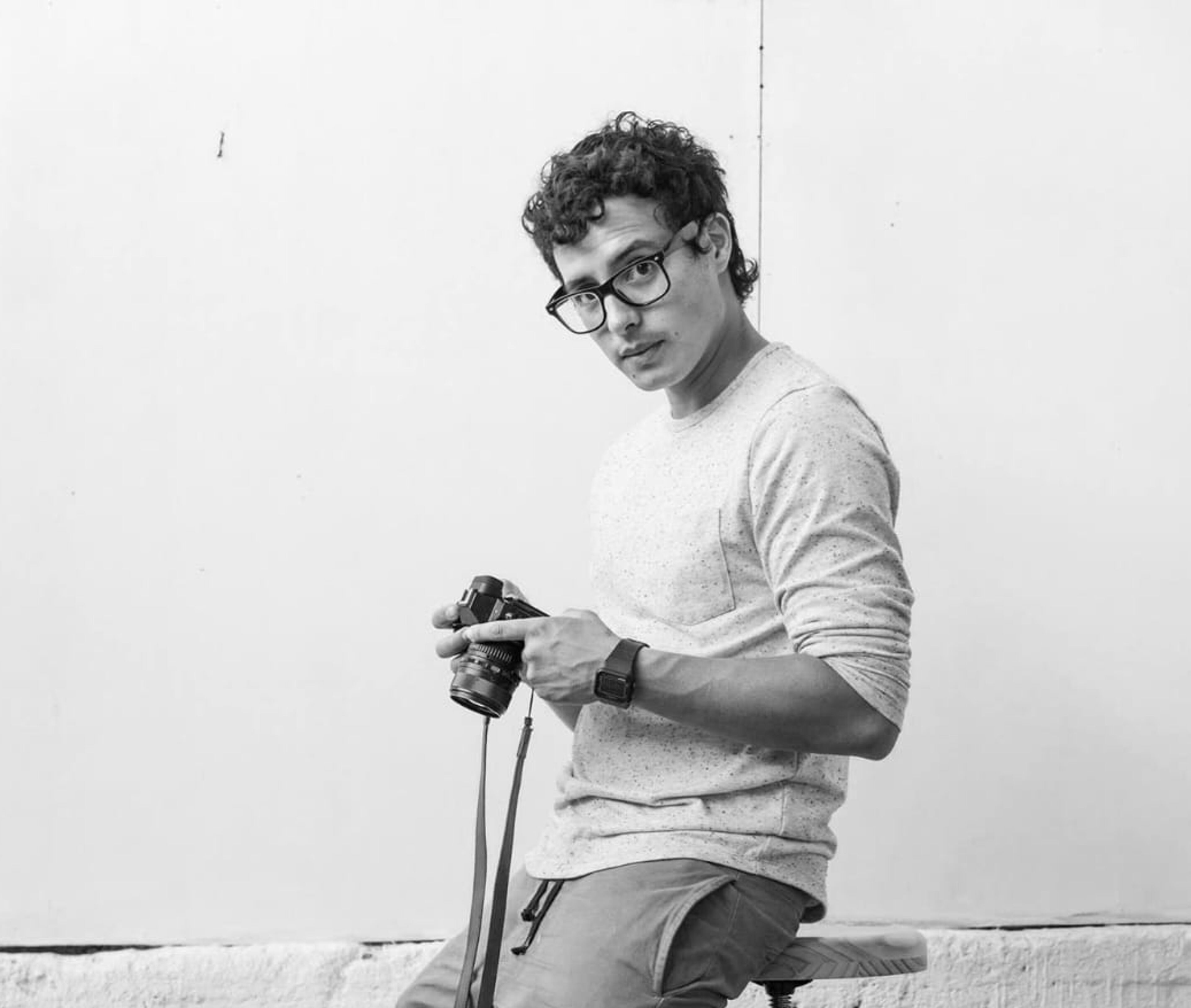 Un joven con pelo rizado y gafas se apoya en un taburete, sosteniendo una cámara, en una foto en blanco y negro.