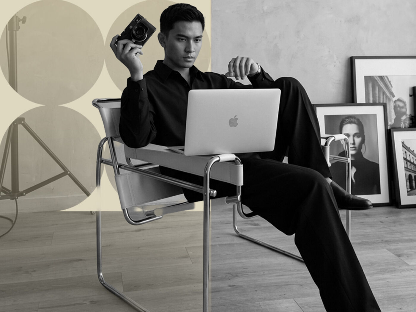 A young man holding a camera and with a laptop on his lap, seated in a studio chair, with framed portraits in the background.