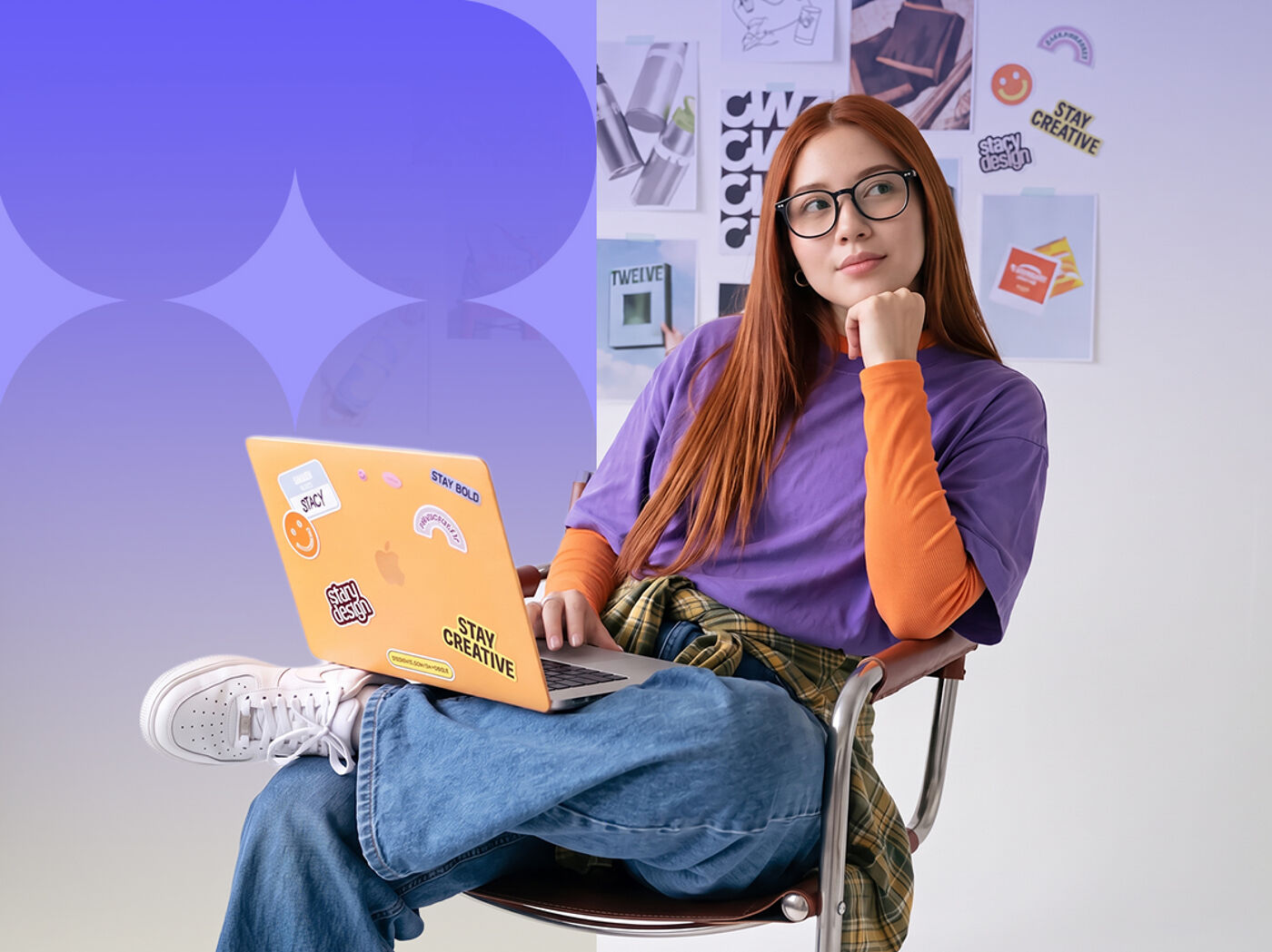 A young woman with red hair and glasses is sitting in a chair with an orange laptop on her lap, looking thoughtful. She has decorated her laptop with stickers.