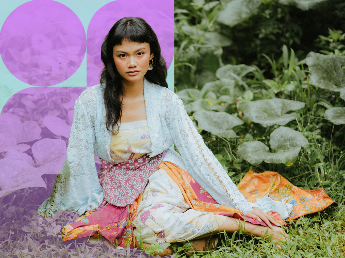 A young woman with dark hair and large earrings sits on the grass wearing a colorful traditional dress and a light cardigan. She is surrounded by green vegetation and large leaves.