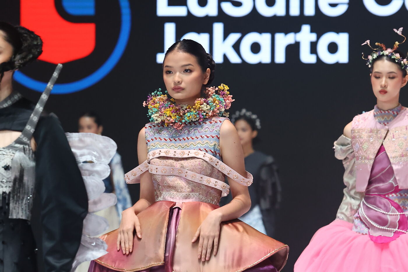 Models showcase avant-garde fashion with intricate designs and colorful accessories on a runway, possibly at a design school event in Jakarta.