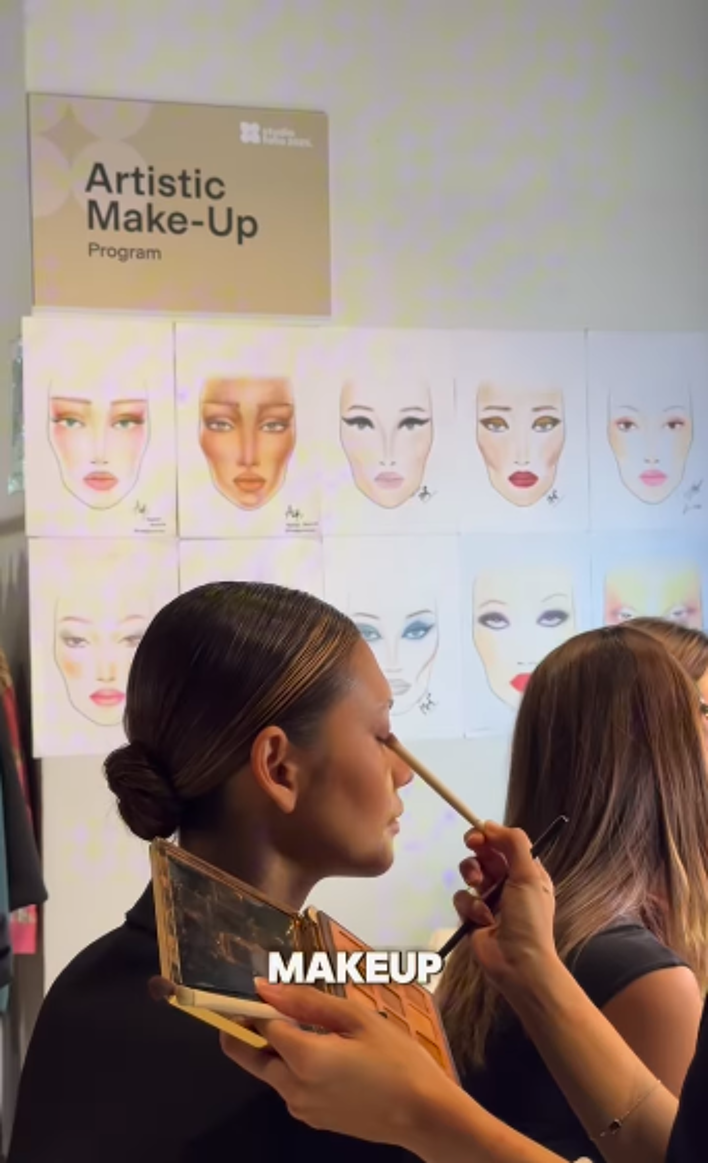A woman is getting her makeup done by a makeup artist. There are several face charts with different makeup looks displayed on the wall behind them. The makeup artist is using a brush to apply eyeshadow to the woman's eyelid.