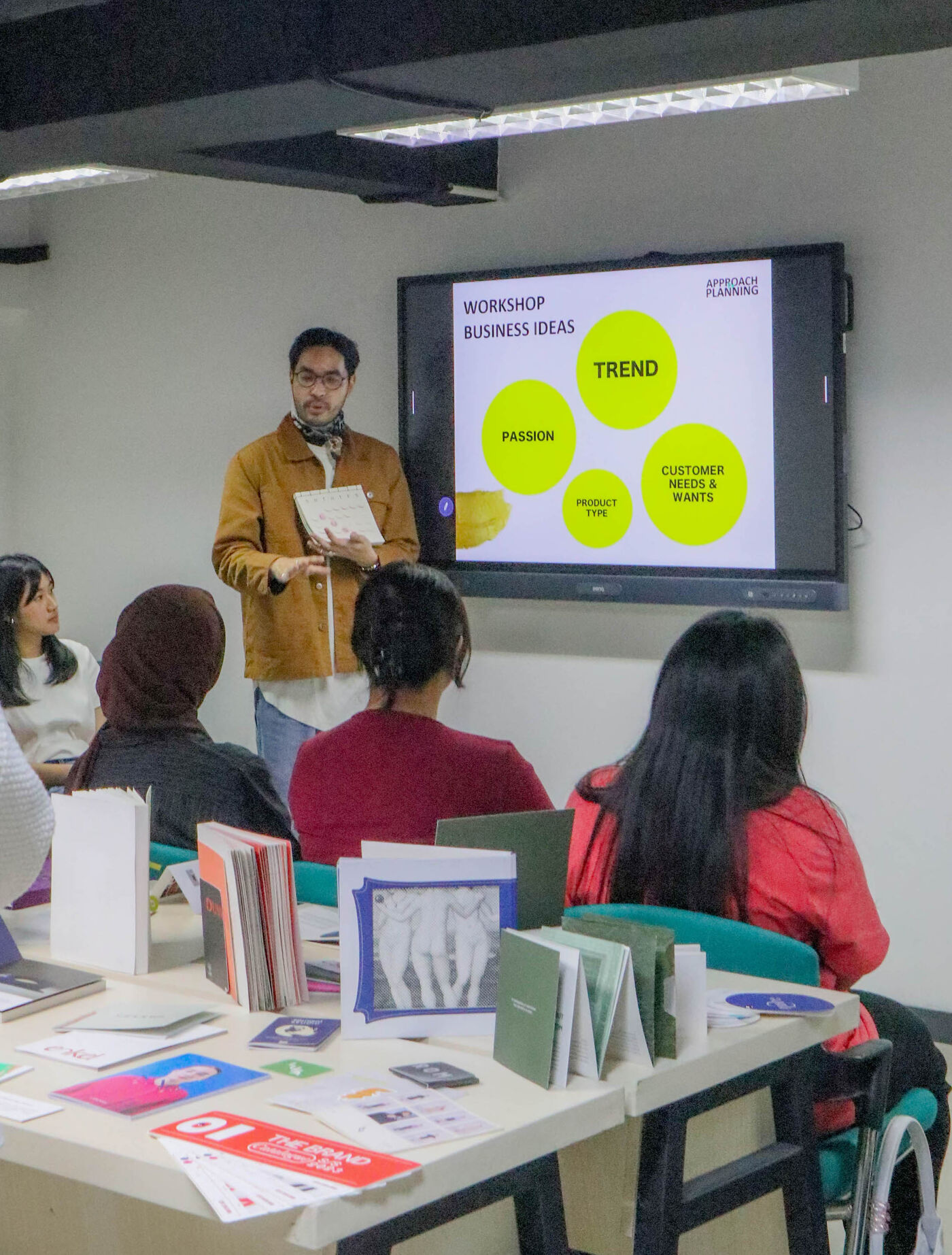 A man presents business ideas in a workshop, with a chart showing trends, passion, and customers. Several people are attending the presentation.