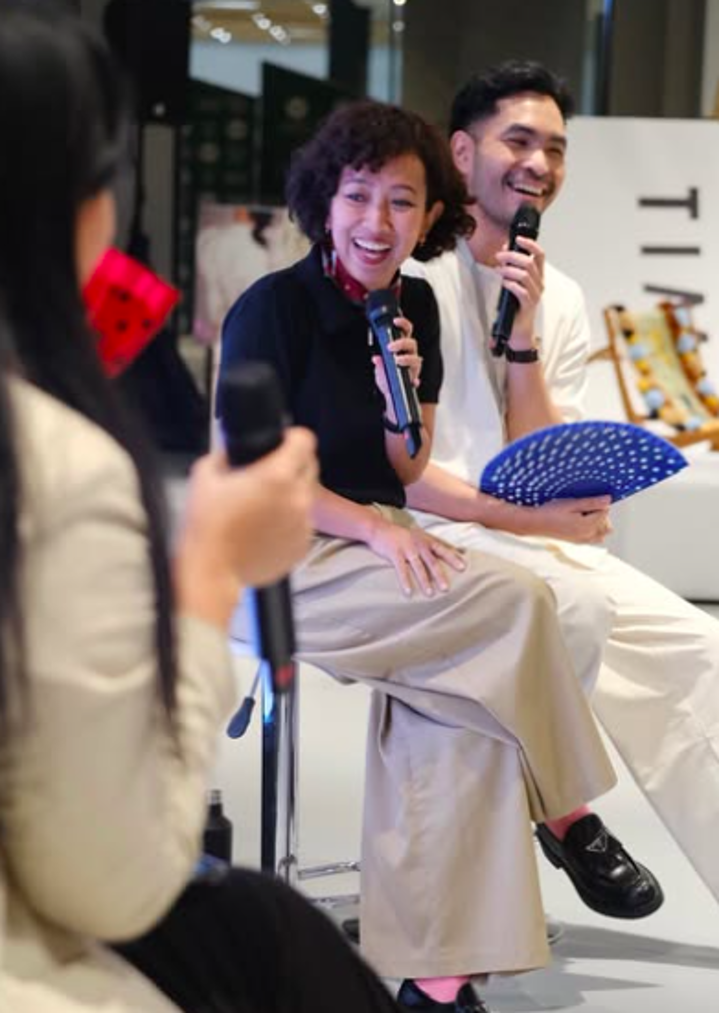 The image shows two people, a woman and a man, sitting on stools and holding microphones, seemingly engaged in a discussion or presentation. The woman is laughing, while the man is smiling. A third person is partially visible on the left.