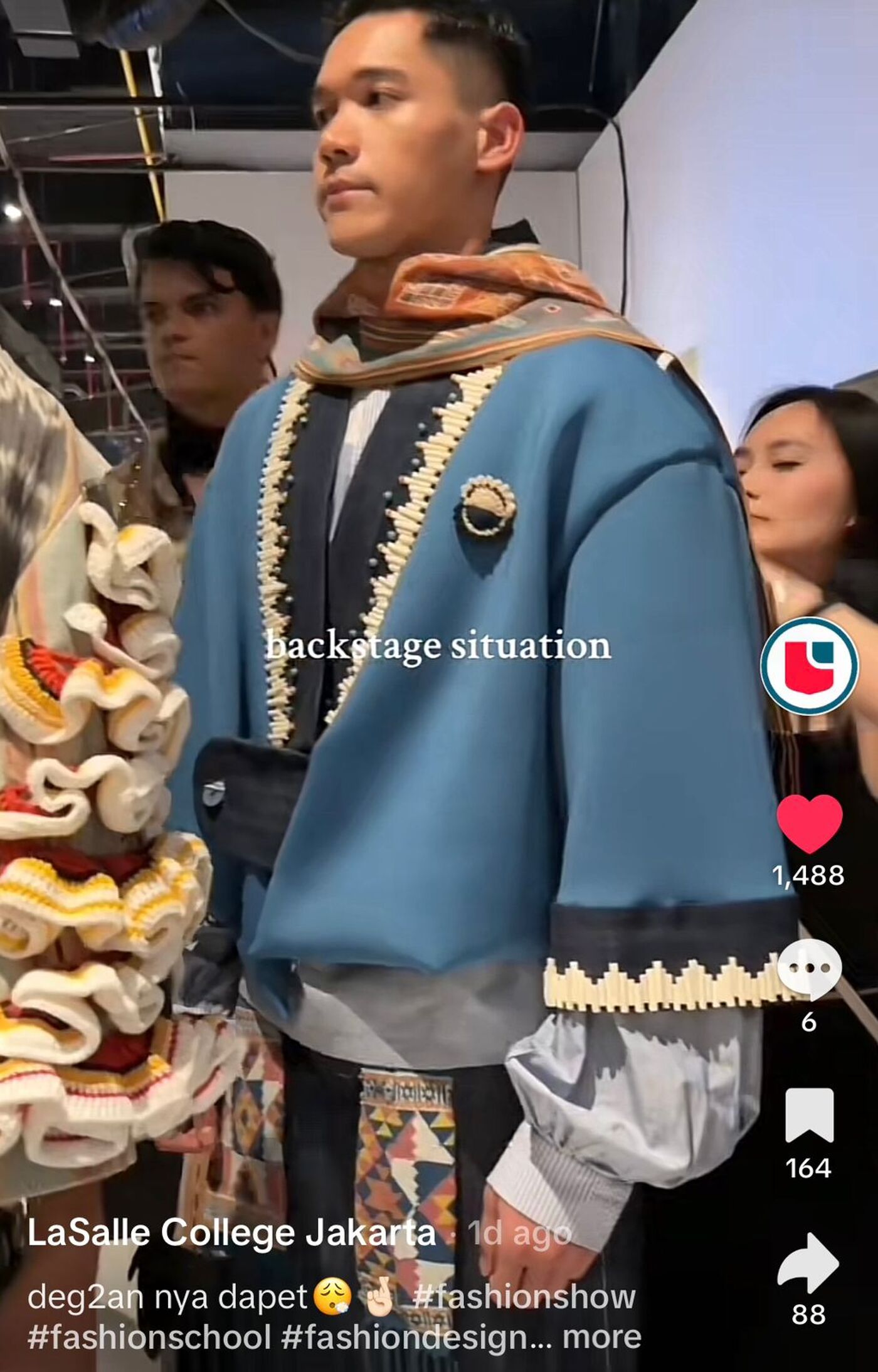 The image shows a person in a blue and black outfit with intricate detailing, likely a fashion design. The person is standing in what appears to be a backstage area, possibly at a fashion show or photoshoot.
