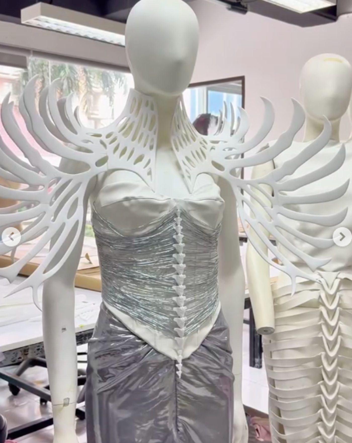 The image shows a mannequin dressed in a futuristic white and silver outfit. The outfit features a structured bodice, a flowing skirt, and a decorative white collar and shoulder piece resembling bones or branches.