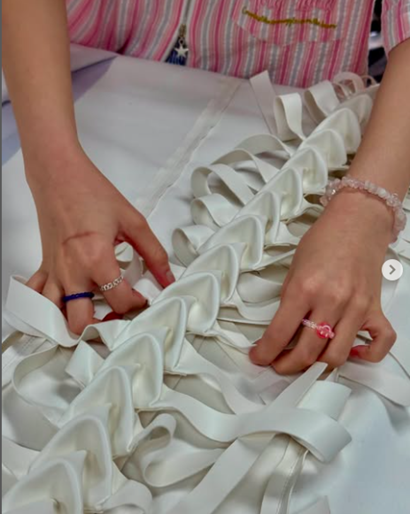 A close-up shot shows a person's hands arranging white satin ribbons on a white fabric base. The ribbons are layered and create a textured, ruffled effect, possibly for a garment or decorative piece.