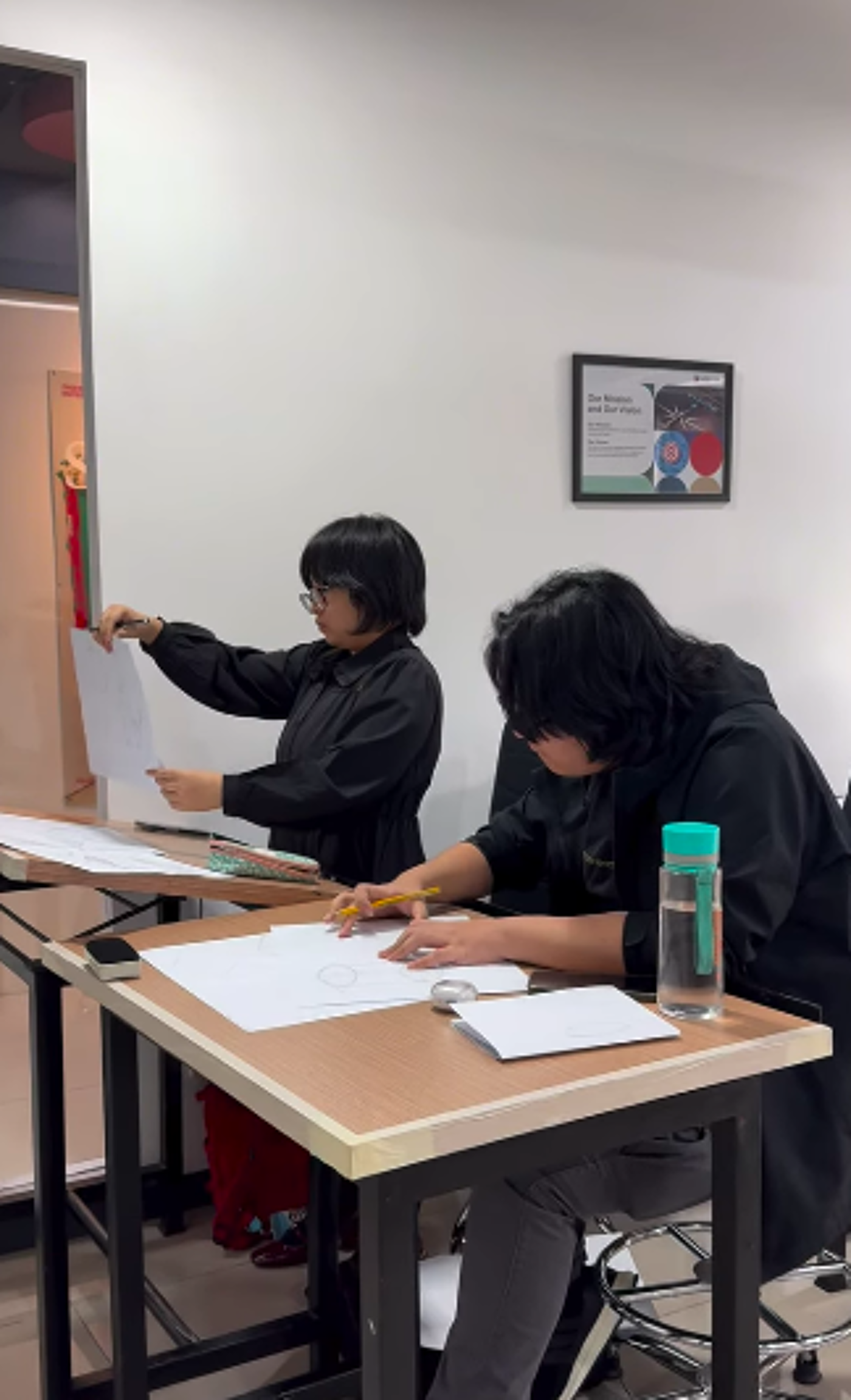 Two people are sitting at desks, seemingly engaged in a drawing or art-related activity. One person is holding up a piece of paper, possibly for reference, while the other is actively drawing on a sheet of paper.
