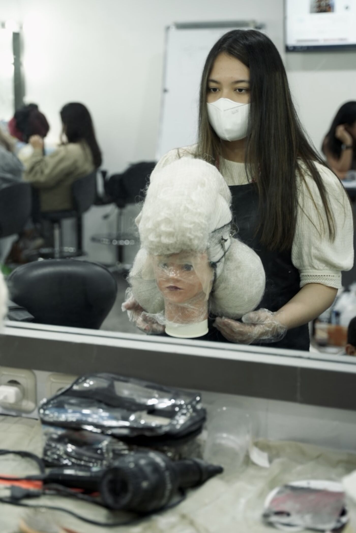 A woman wearing a mask holds a mannequin head with curly white hair, possibly in a hairstyling or cosmetology class.