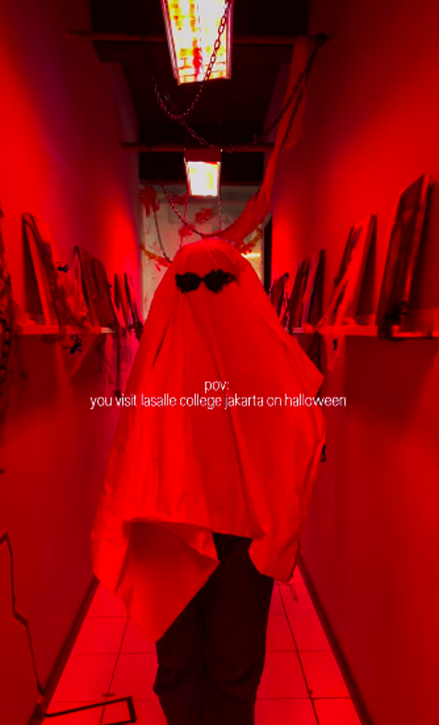 The image shows a person dressed as a ghost in a hallway, likely for Halloween at Lasalle College Jakarta. The scene is bathed in red light, creating a spooky atmosphere, with decorations like spiderwebs and chains enhancing the Halloween theme.