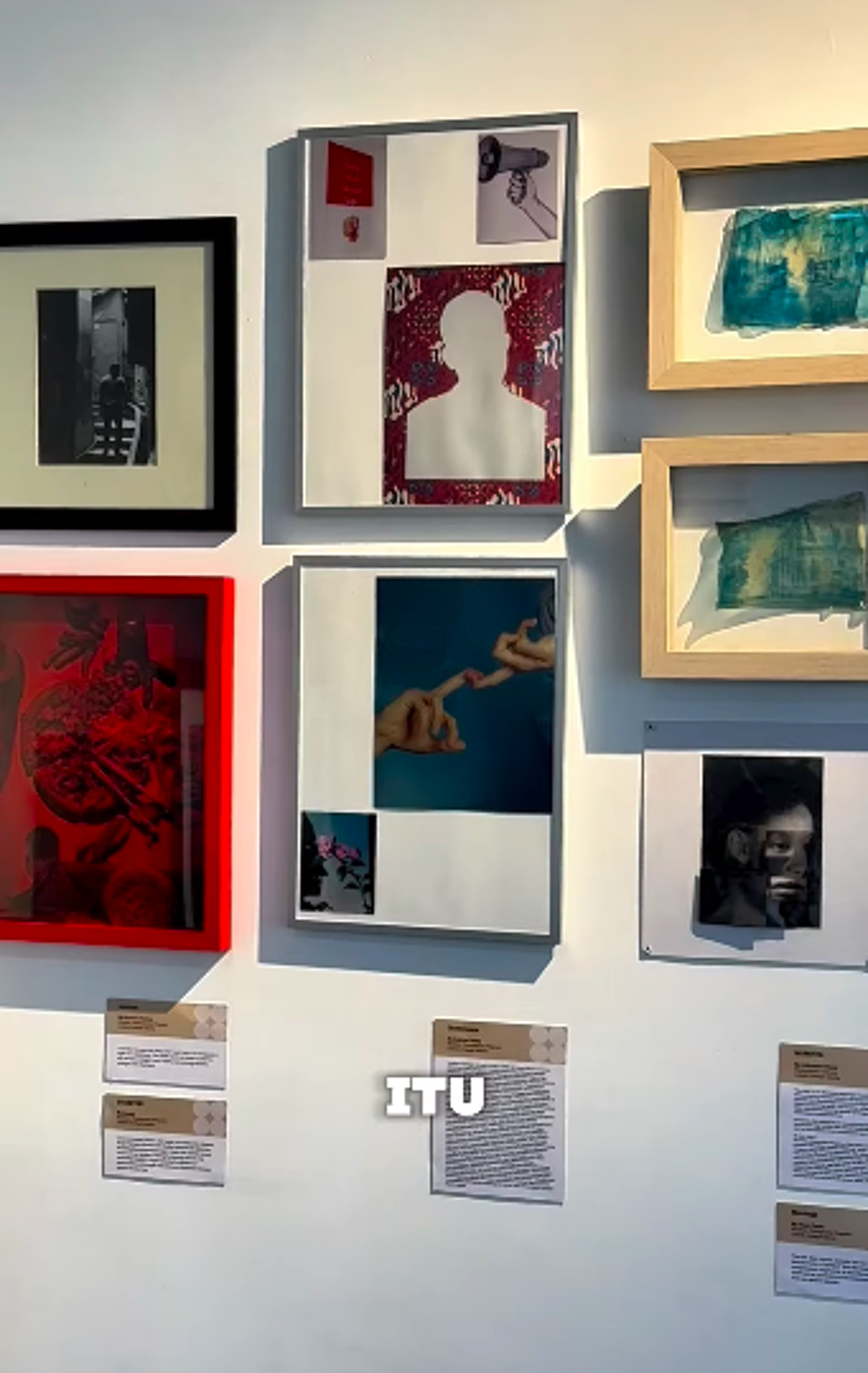 The image shows a wall in an art gallery with several framed pieces of art. The artworks are diverse, including photography, collages, and abstract pieces, each with accompanying descriptions below.