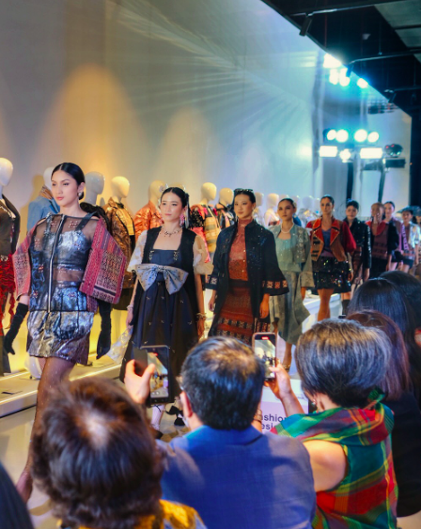 A fashion show is underway with models walking down a runway showcasing various designs. Attendees are capturing the event with their phones. The designs appear to incorporate traditional patterns and modern styles.