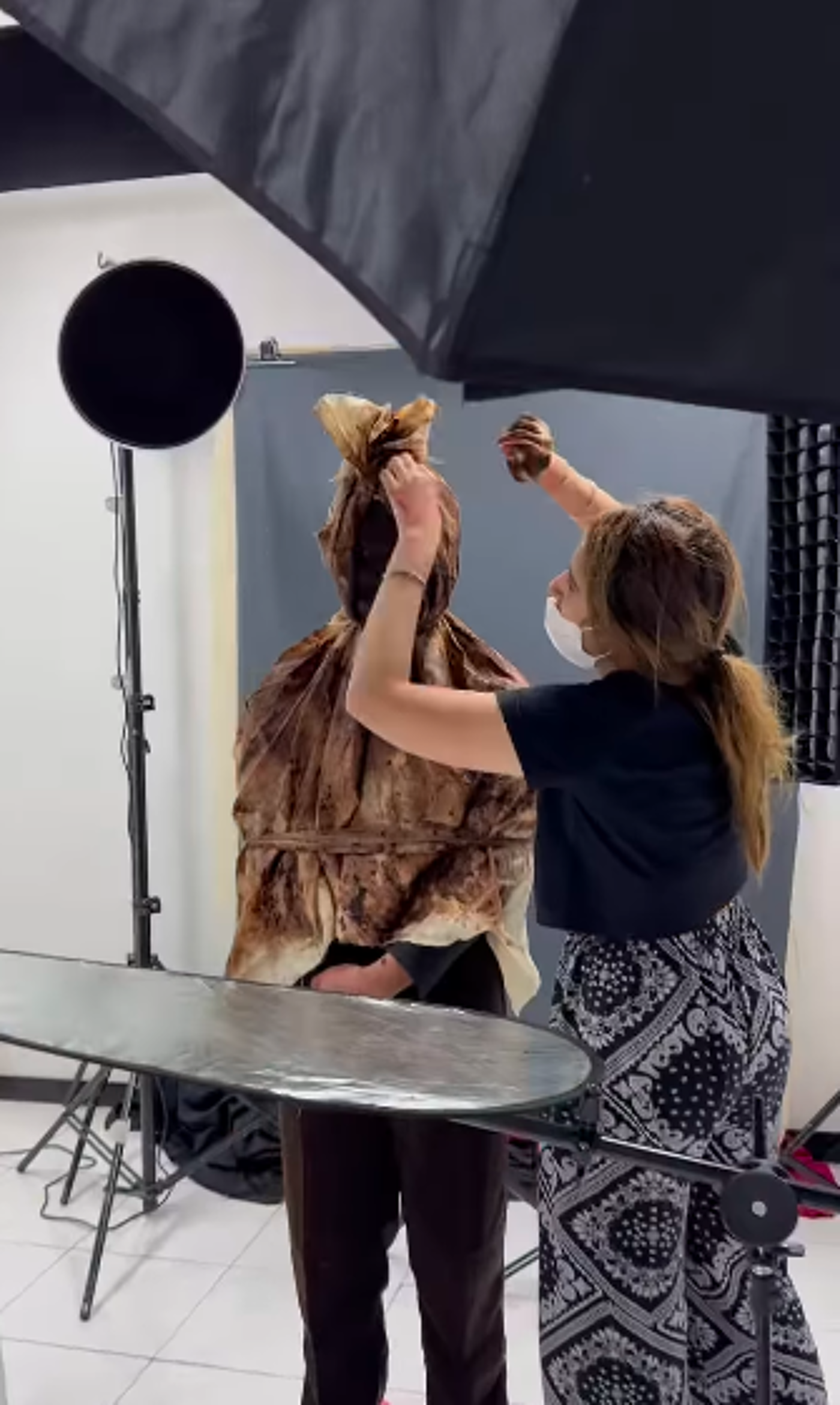 A person is being styled in a studio, wrapped in brown fabric while a makeup artist adjusts their hair and clothing.
