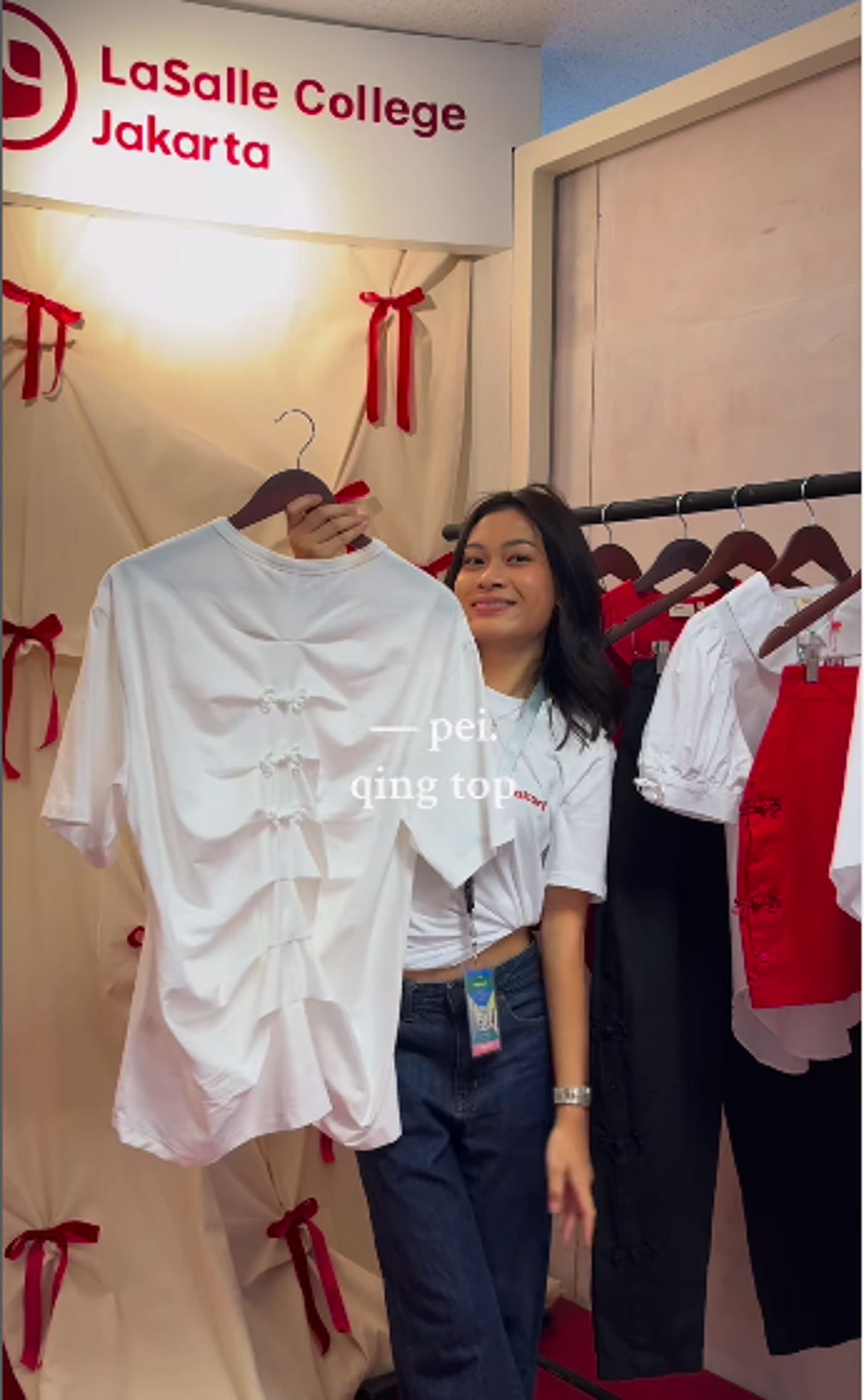 A young woman stands in front of a La Salle College Jakarta sign, holding up a white shirt on a hanger. She is smiling and wearing jeans and a white t-shirt. There are clothes hanging on a rack behind her.