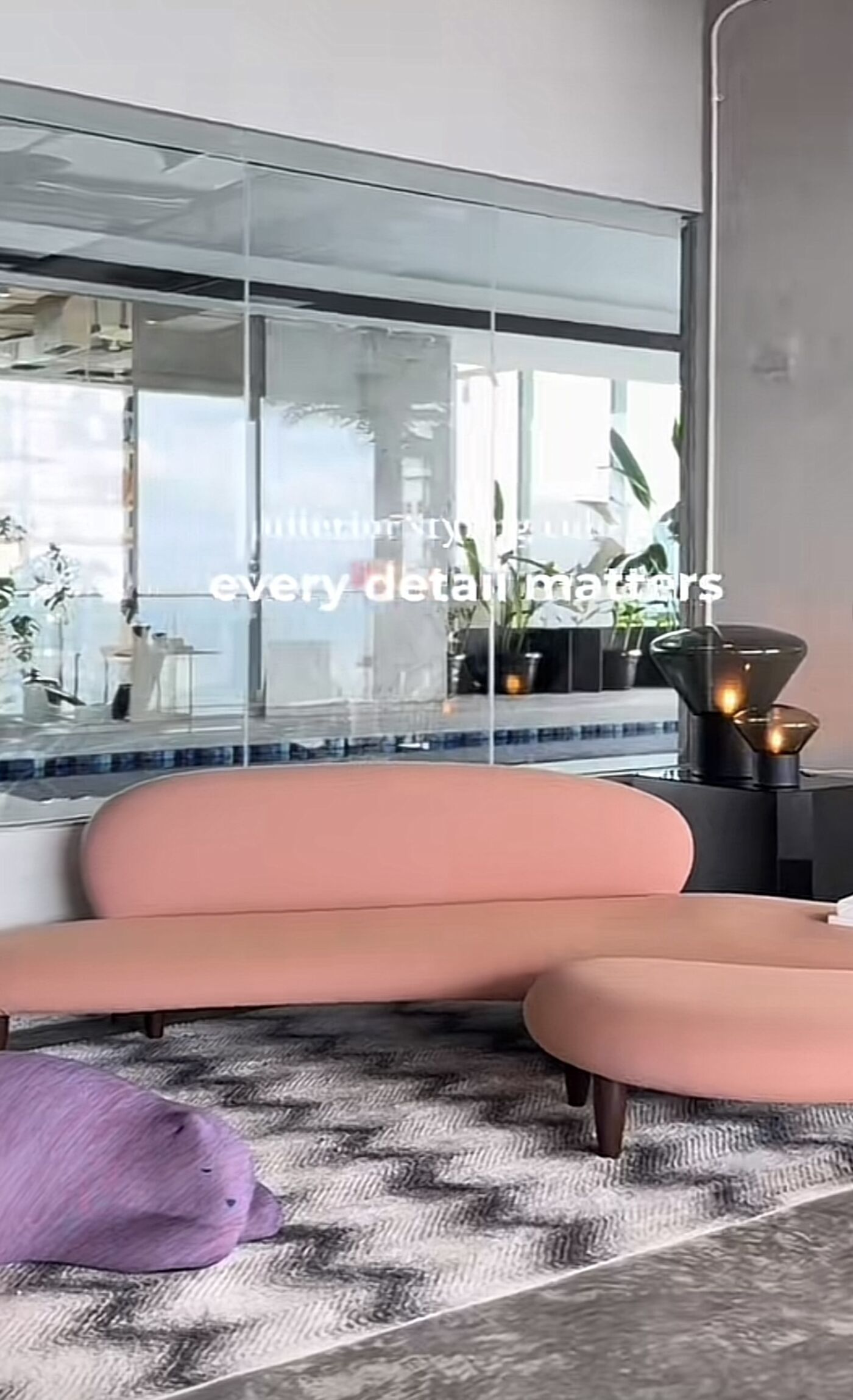 The image shows a modern interior with a pink sofa and ottoman on a patterned rug. A glass wall reflects an outdoor scene with water and greenery. Decorative lamps and a purple pillow add to the stylish ambiance.
