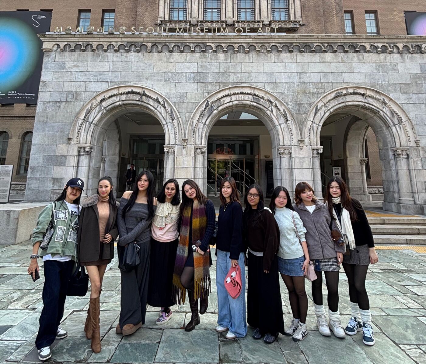 A group of ten young women stand in front of a historic building with arches, dressed in outerwear, posing for a photo on a clear day.