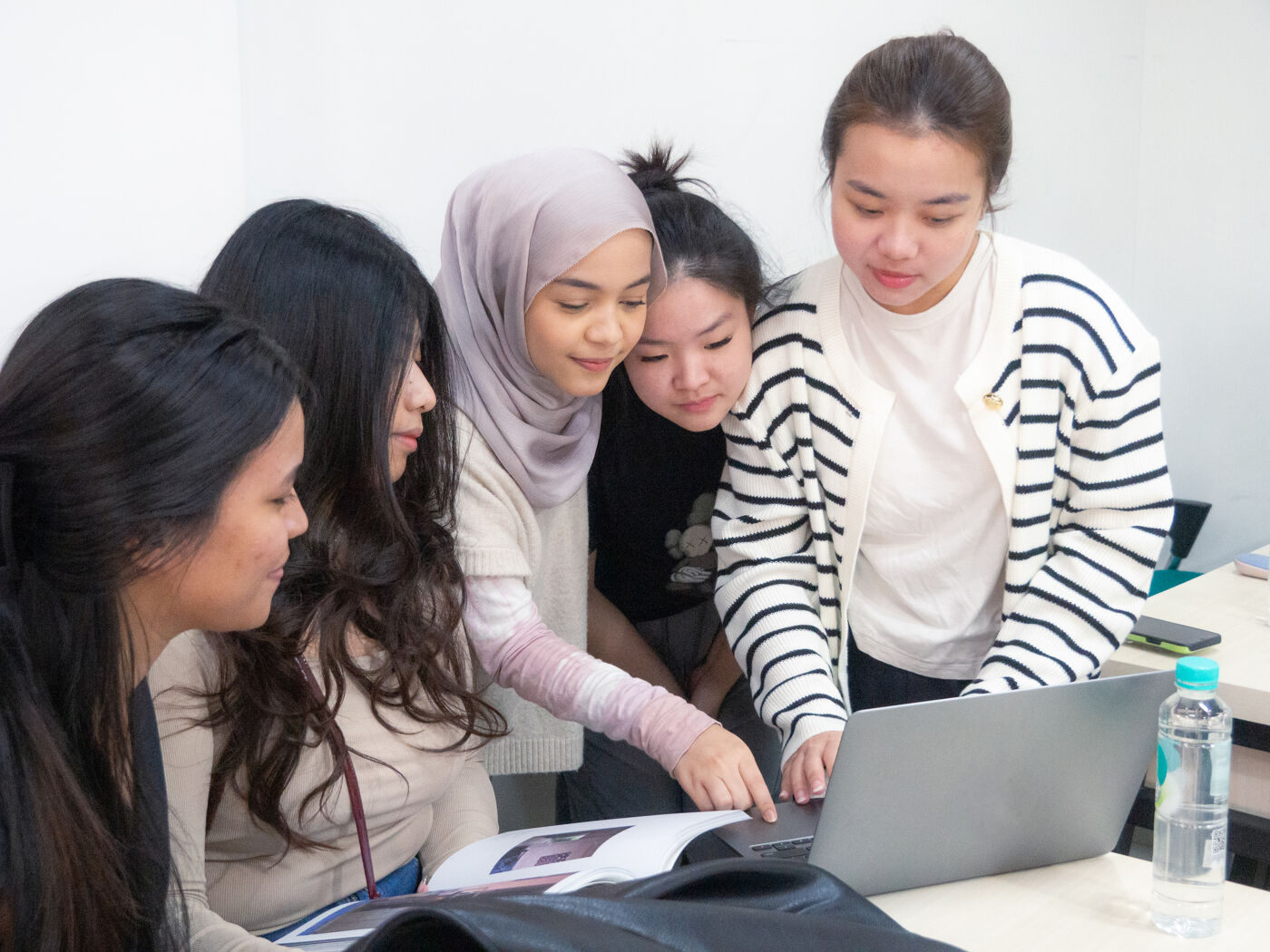 A group of five young women, including one wearing a hijab, attentively focused on a laptop screen, collaborating on a project in a classroom setting.