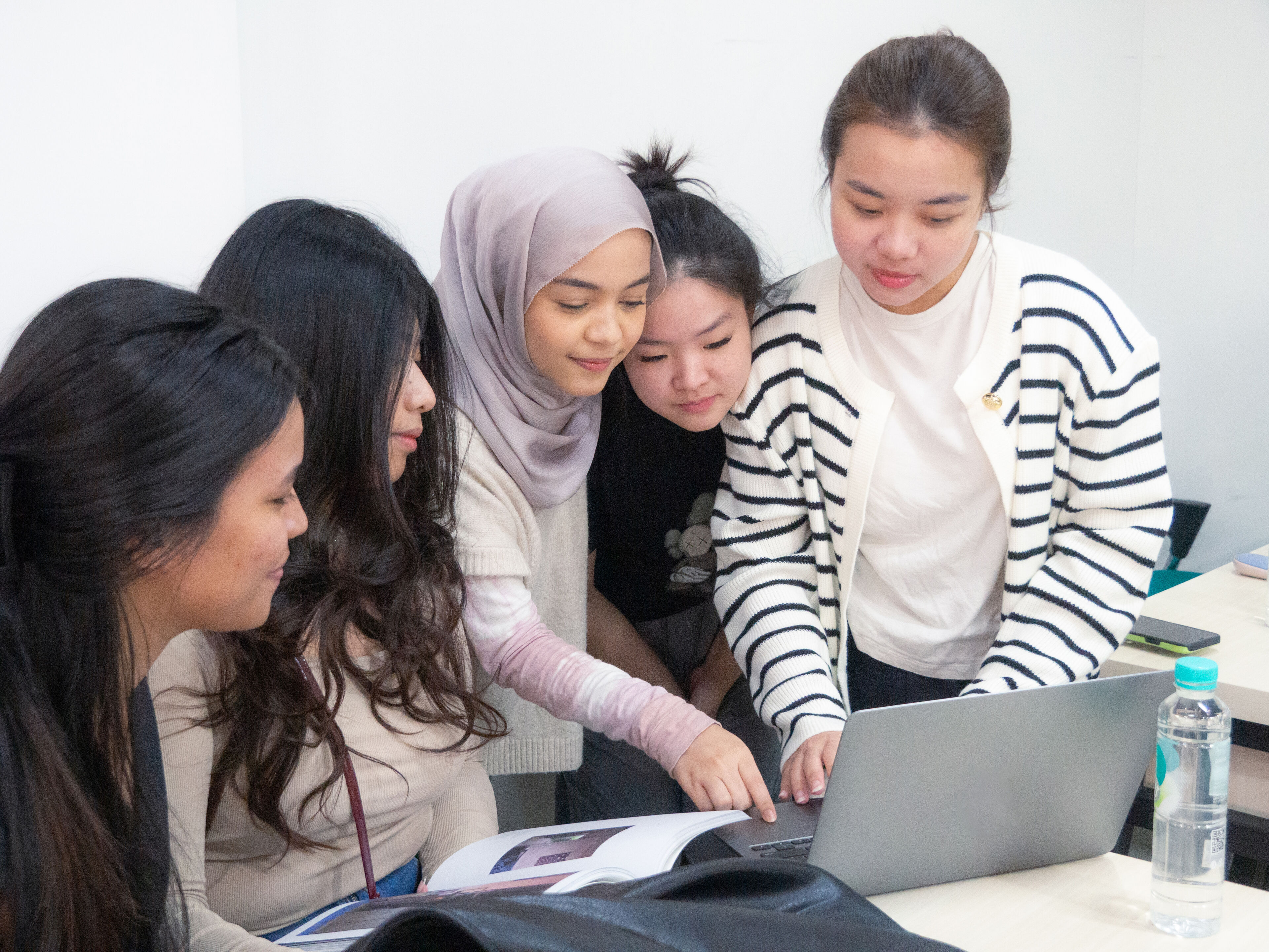A group of five young women, including one wearing a hijab, attentively focused on a laptop screen, collaborating on a project in a classroom setting.
