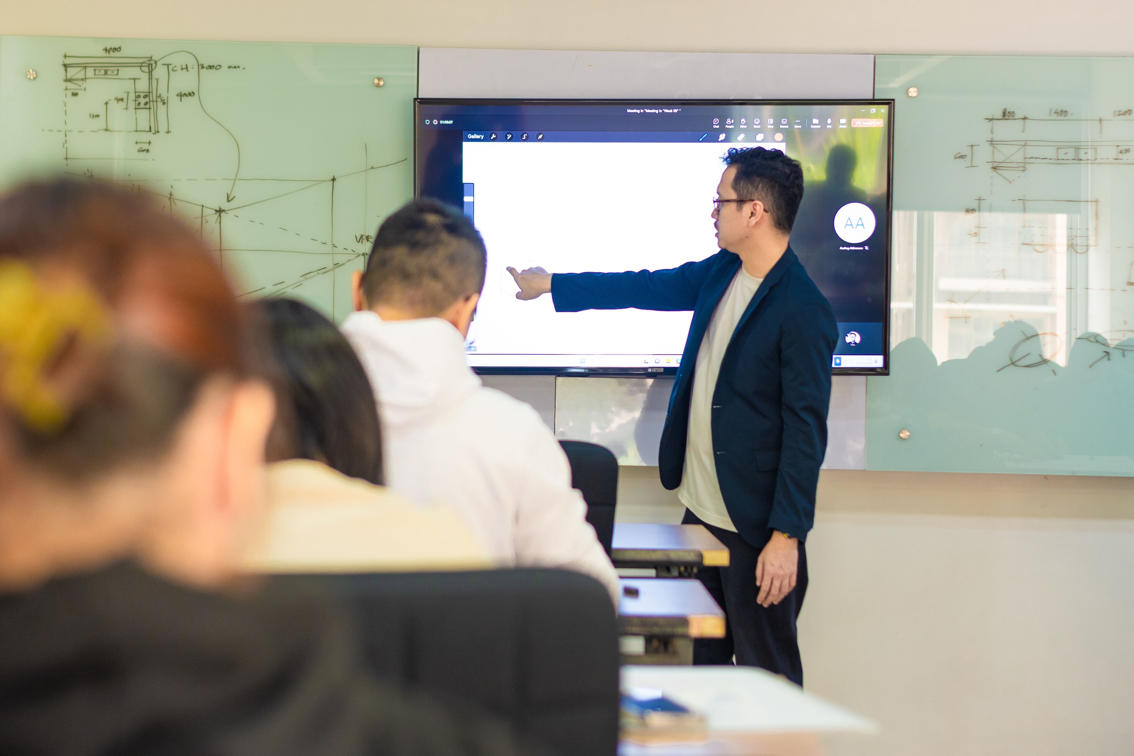 A lecturer pointing to a projection screen with technical drawings, in a classroom with attentive students.
