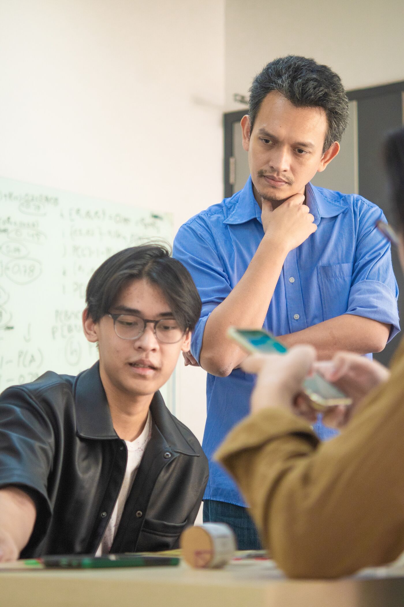 Two male students engage in a thoughtful classroom discussion, one standing with a pensive expression and the other seated, listening attentively.