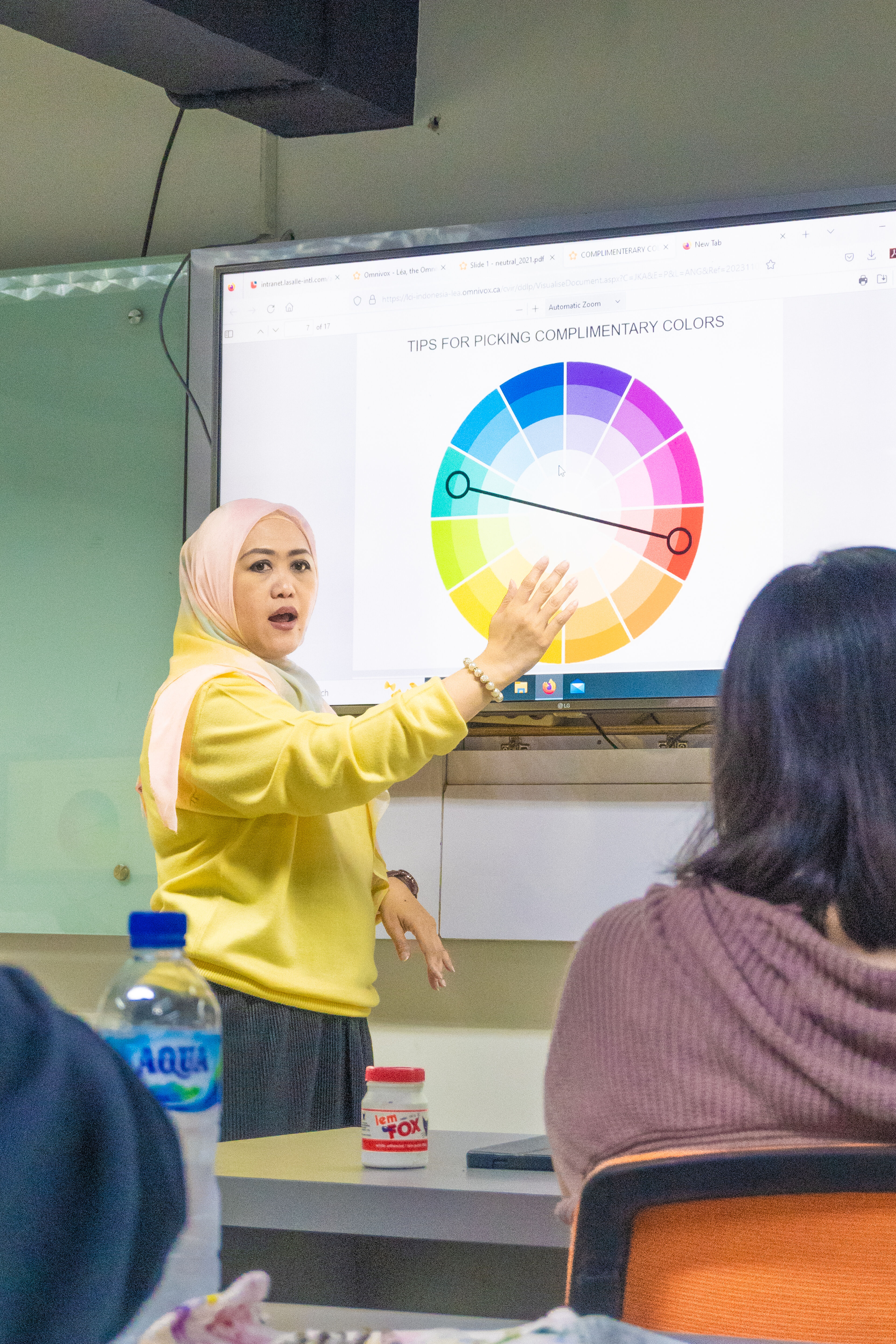 A woman in a hijab presents about complementary colors in a workshop, with a color wheel on the screen.