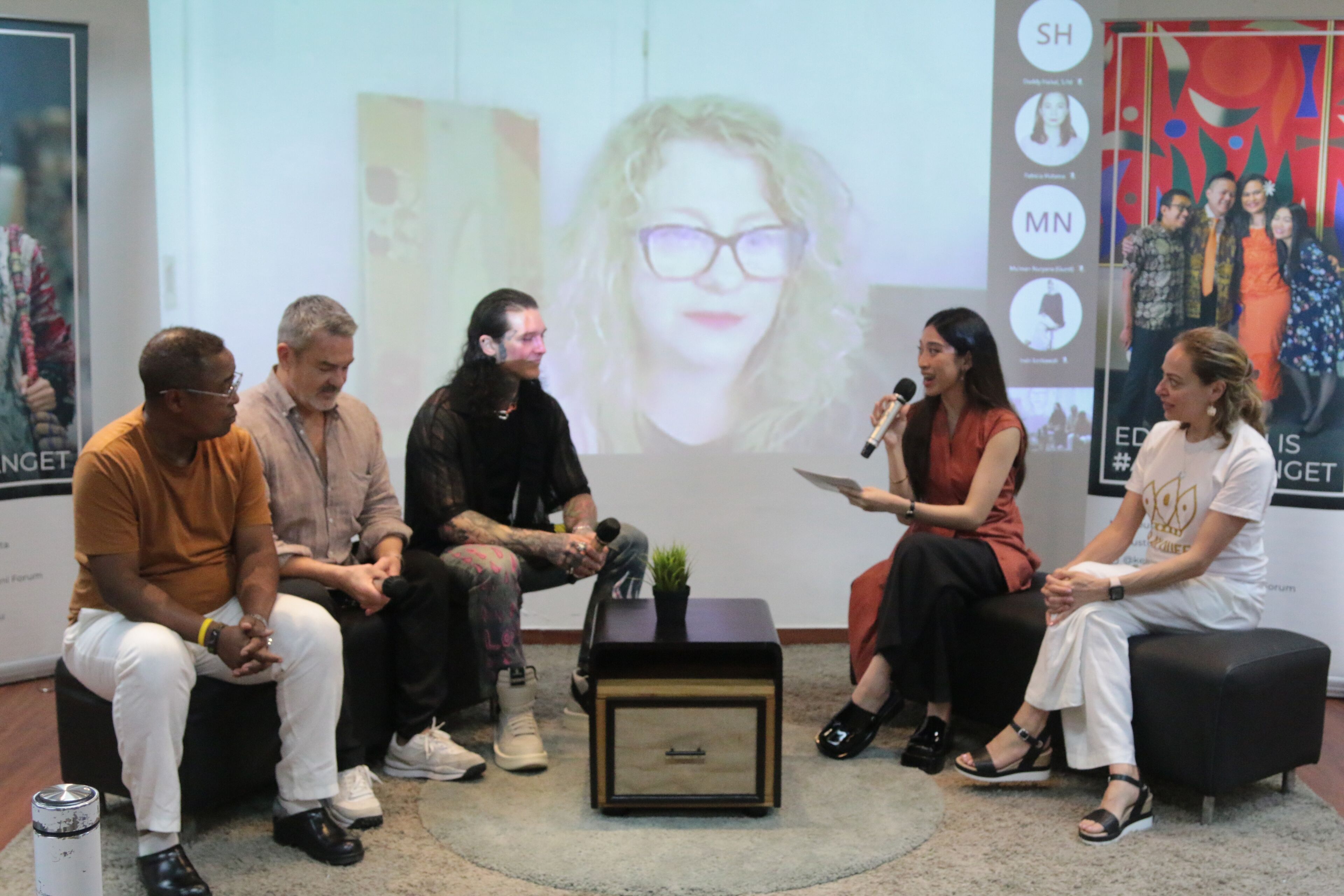 A diverse panel engages in a discussion at an event, including a virtual participant projected on the screen behind them.
