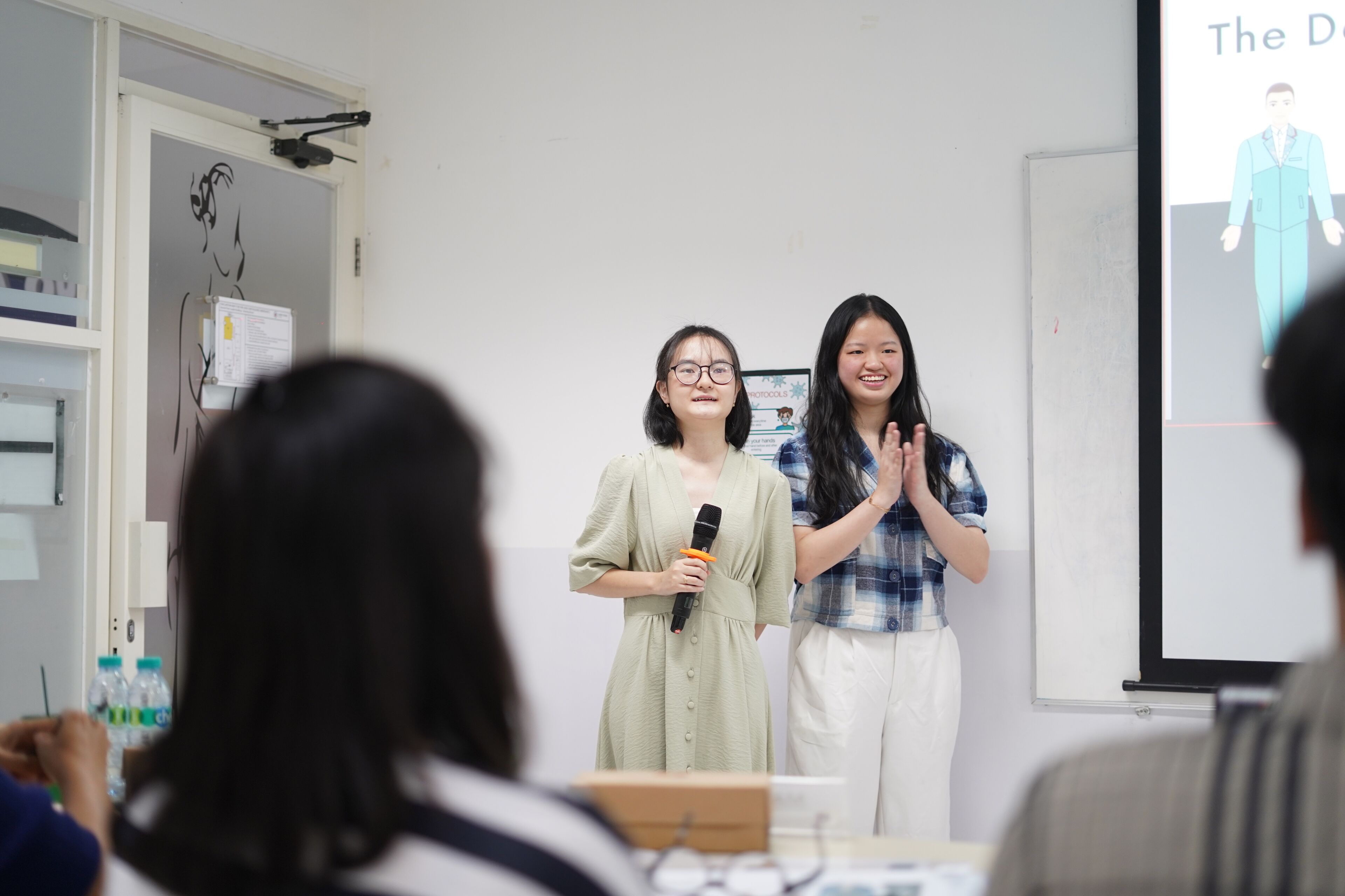 Two students happily present their project in a classroom, one holding a microphone, as the other applauds next to a projected display.