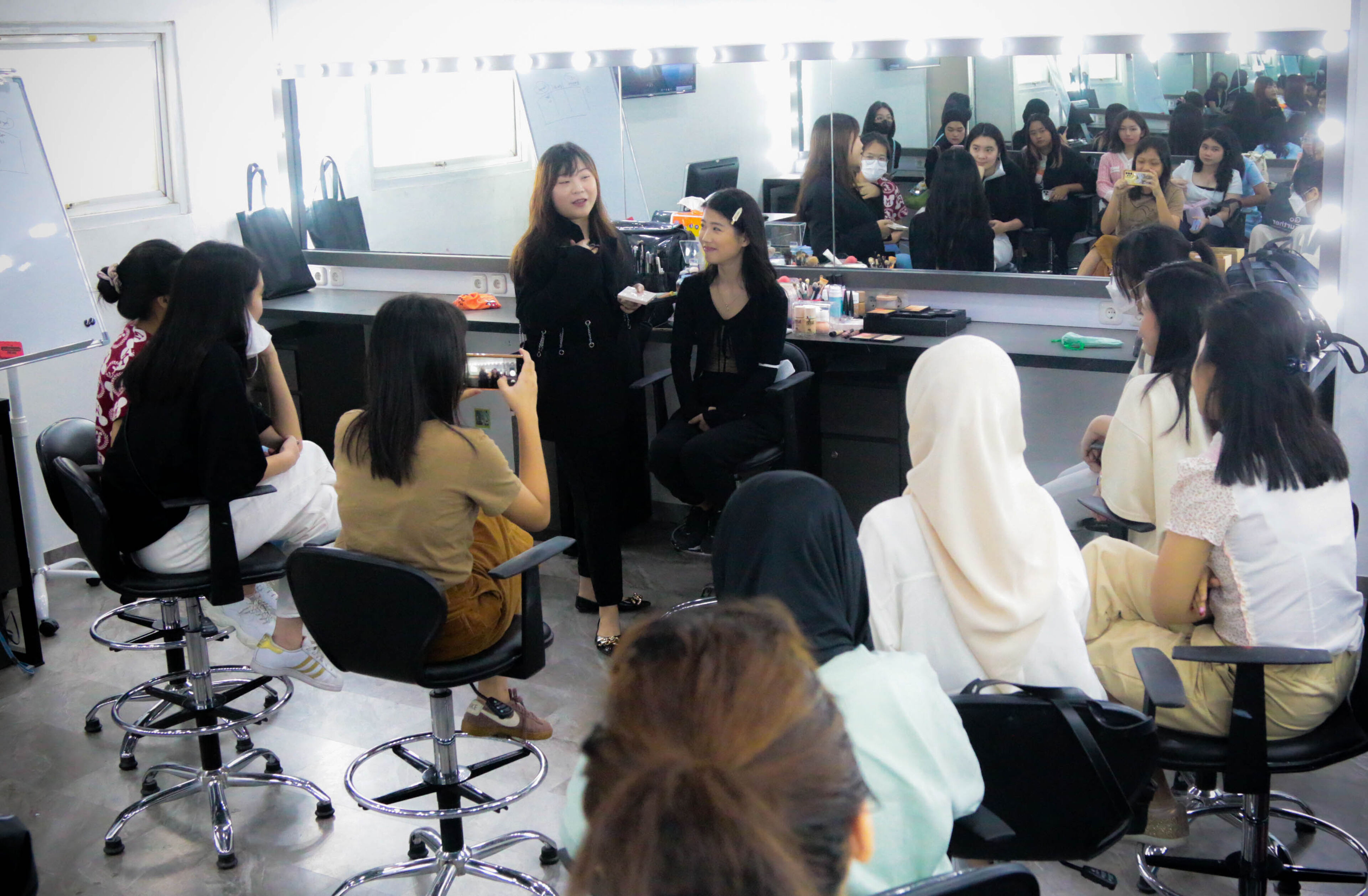 An instructor demonstrates hairstyling techniques to an attentive group of students in a beauty school salon setting, with mirrors reflecting the session.