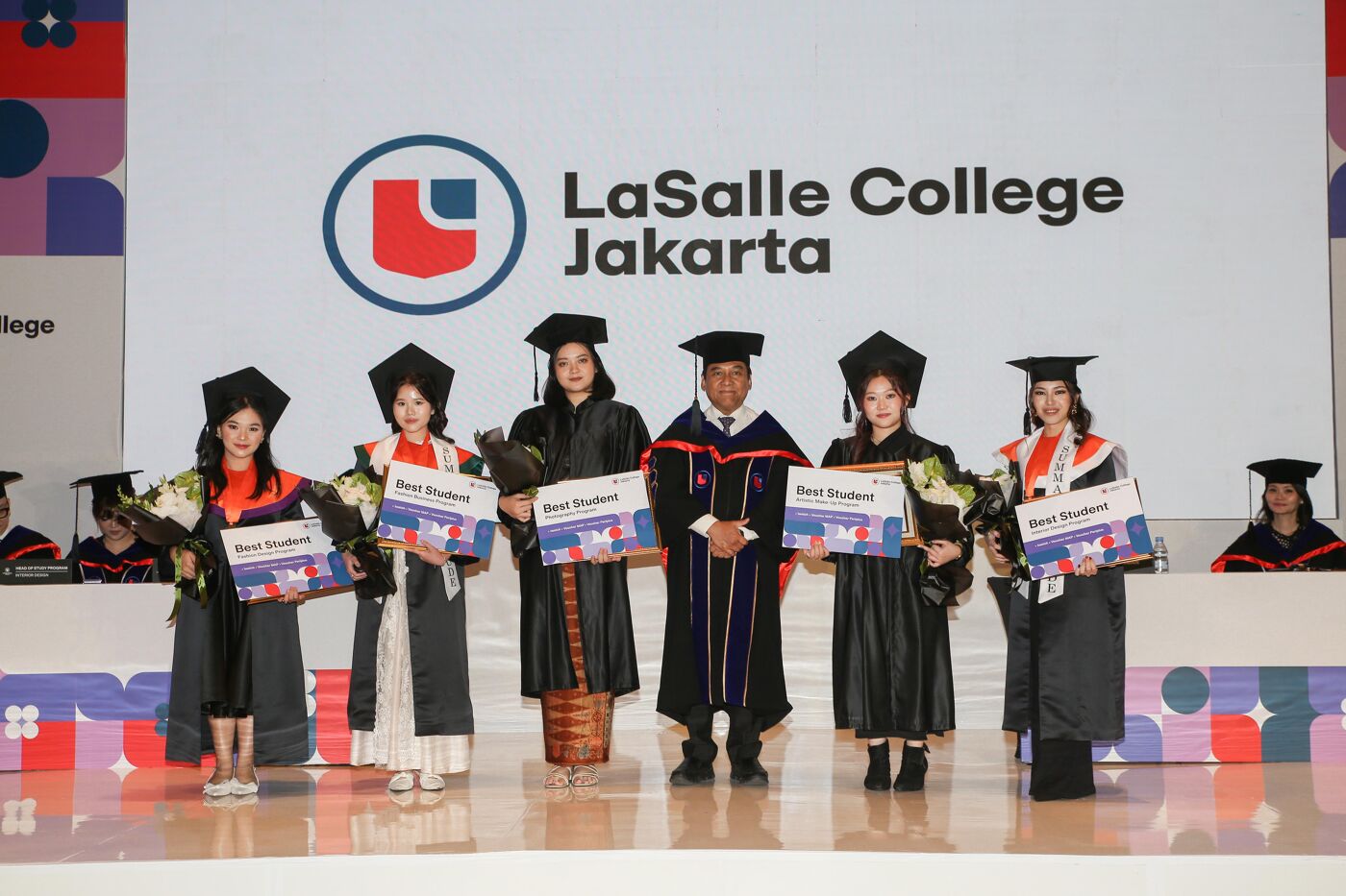 Five graduates proudly hold "Best Student" certificates and bouquets during the graduation ceremony at LaSalle College Jakarta, accompanied by a faculty member in academic regalia. The event backdrop prominently displays the college's logo.