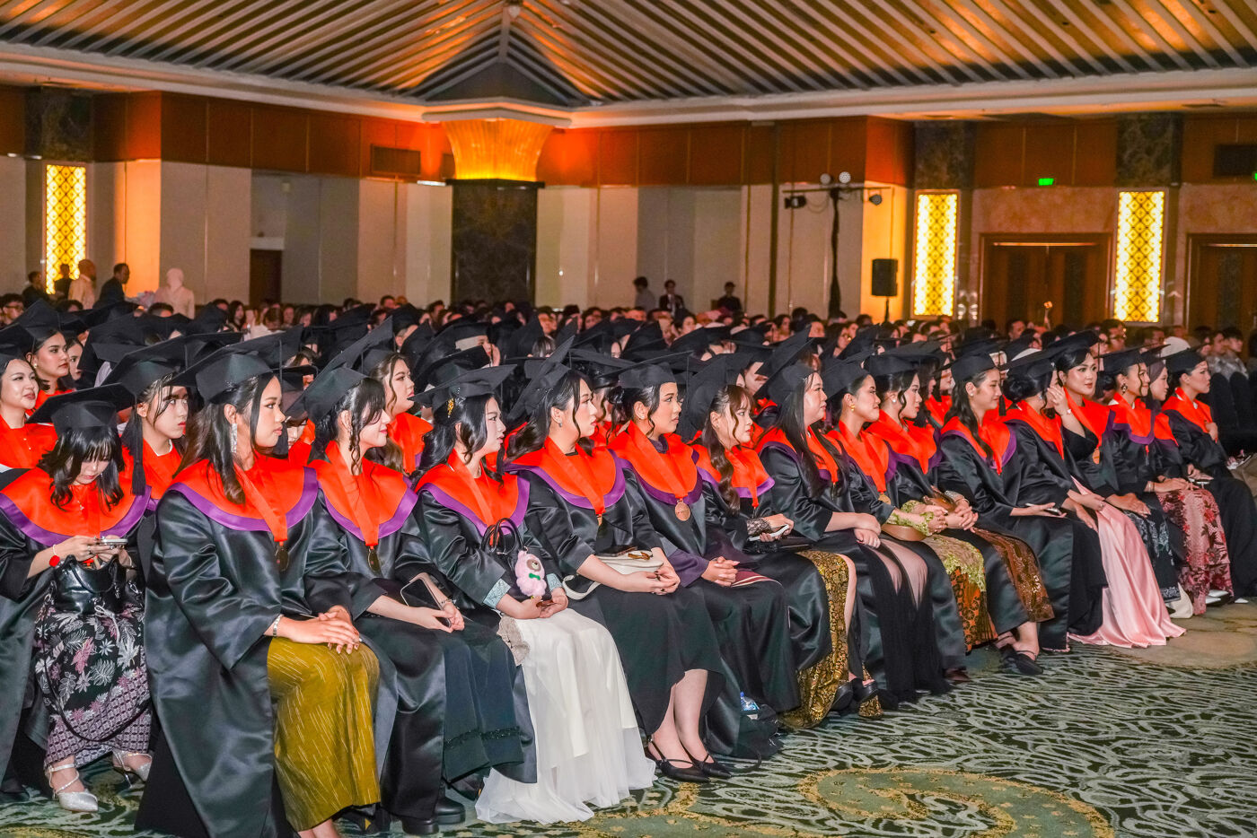 A crowd of graduates wearing gowns and caps are attending a graduation ceremony in a large hall.