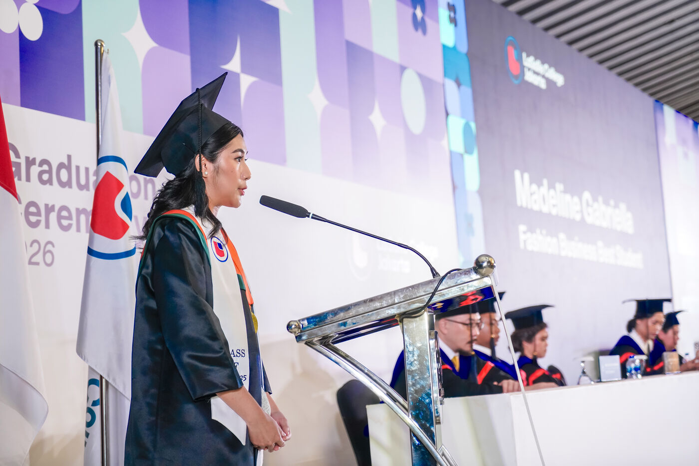 A female graduate speaks at a podium in front of an audience, with a microphone in front of her. Other graduates are seated behind her.