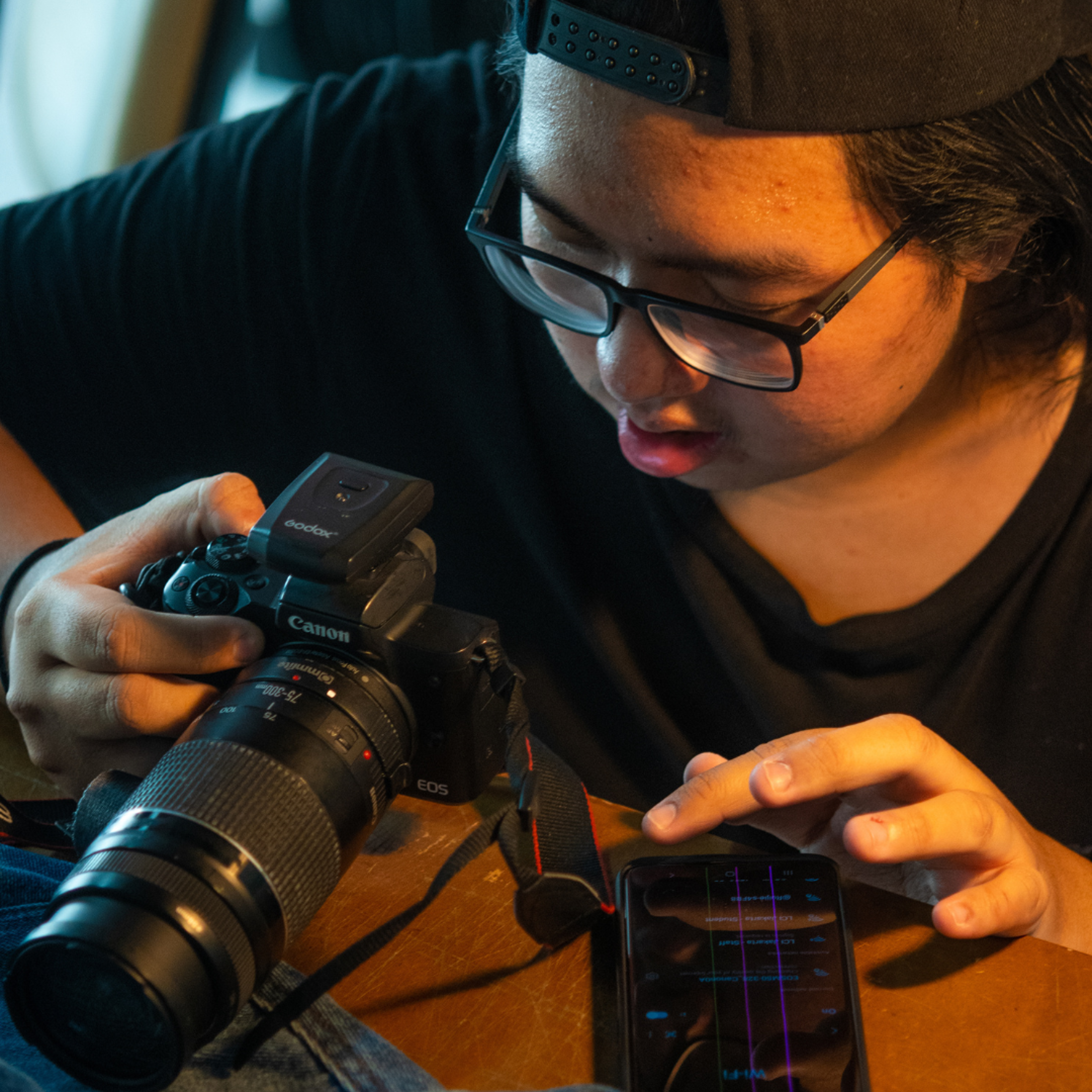 A focused individual adjusts a DSLR camera, with a lens cap and a smartphone displaying graphs placed on a wooden surface nearby.