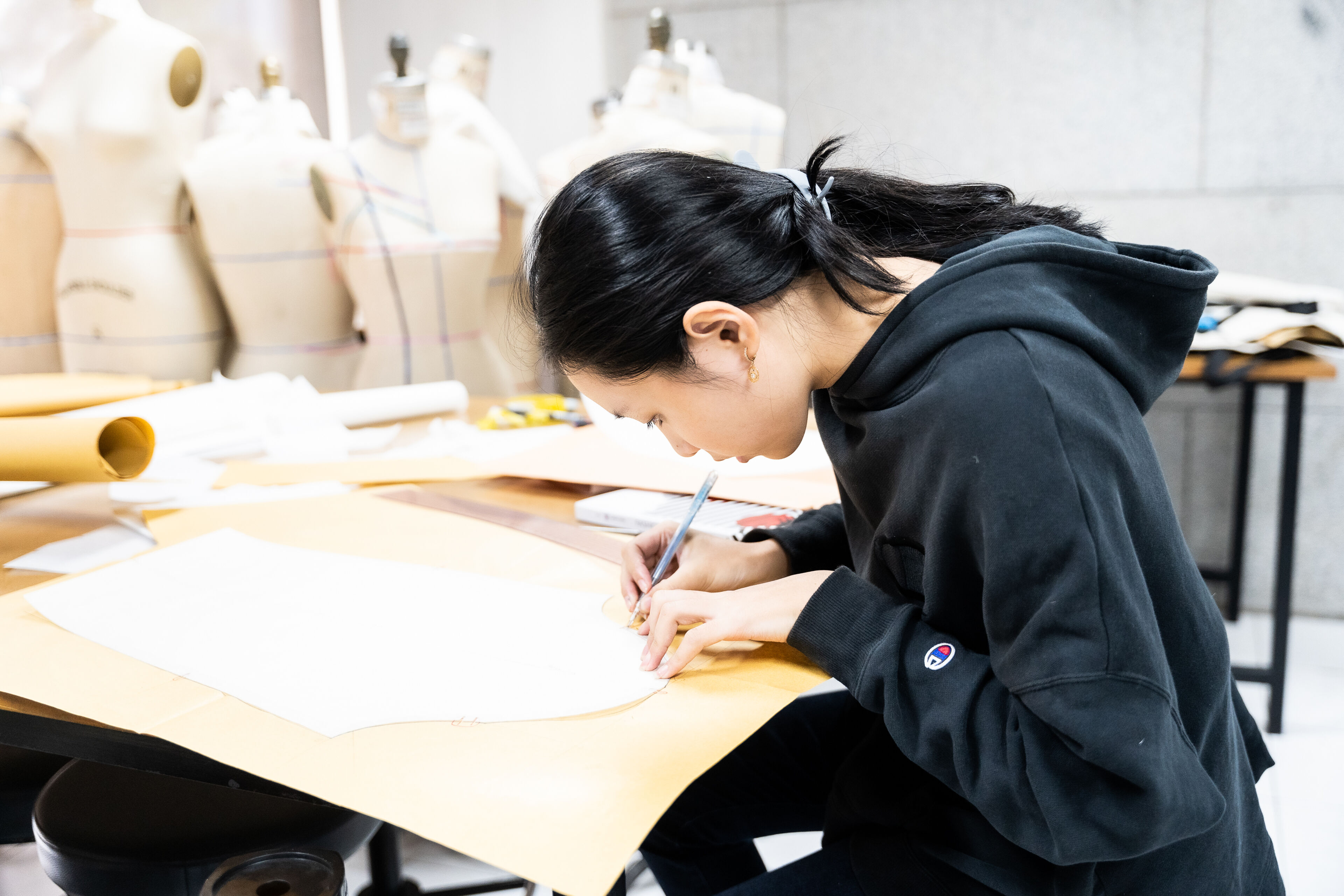A focused fashion designer is meticulously drafting clothing patterns on a large paper at a well-lit workshop table, with mannequins and design tools in the background.