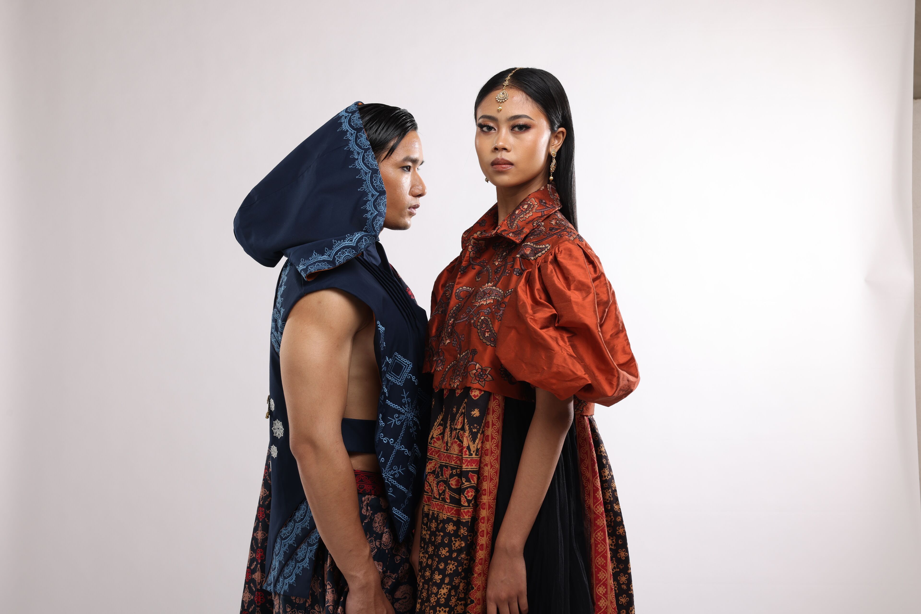 Two models showcase a blend of traditional and modern attire, with one in a blue embroidered hood and the other in an ornate, rust-colored blouse, both exuding cultural richness.