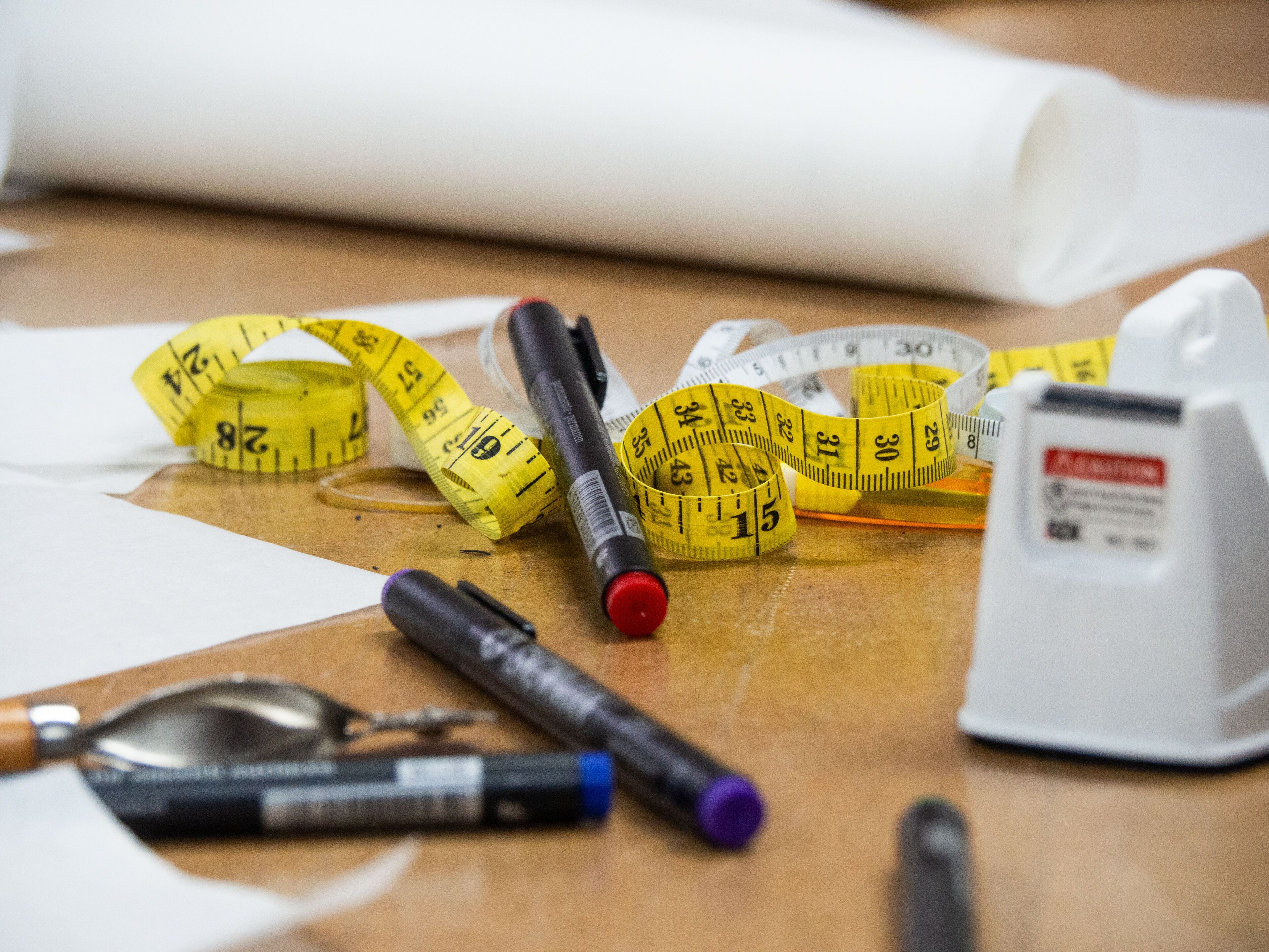 Various tailoring tools, including measuring tapes, markers, and scissors, arranged on a wooden worktable with rolls of paper in the background.