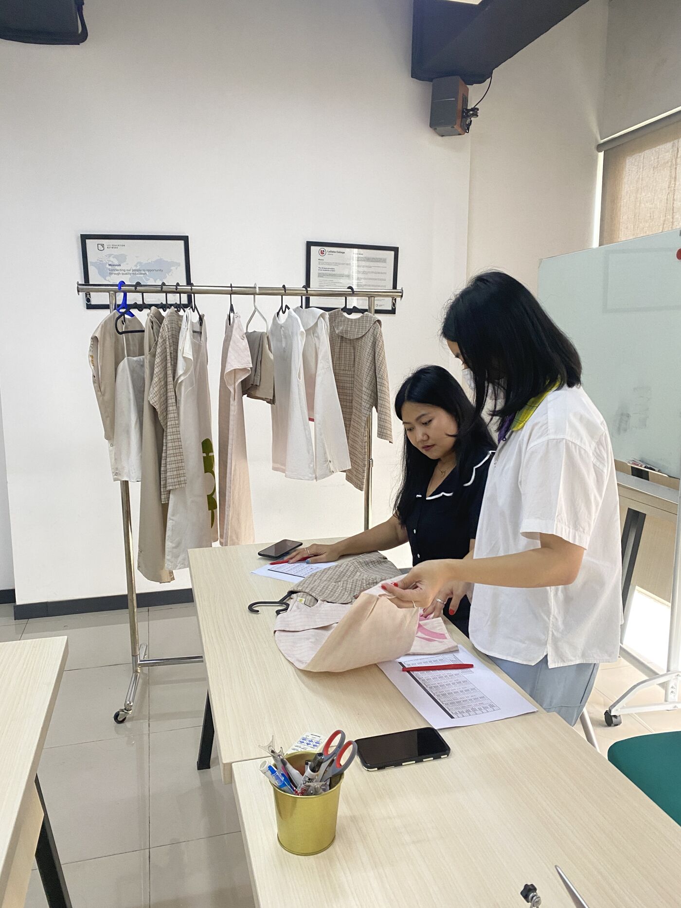 Two women in a workspace, examining a piece of fabric with garments on a rack in the background.