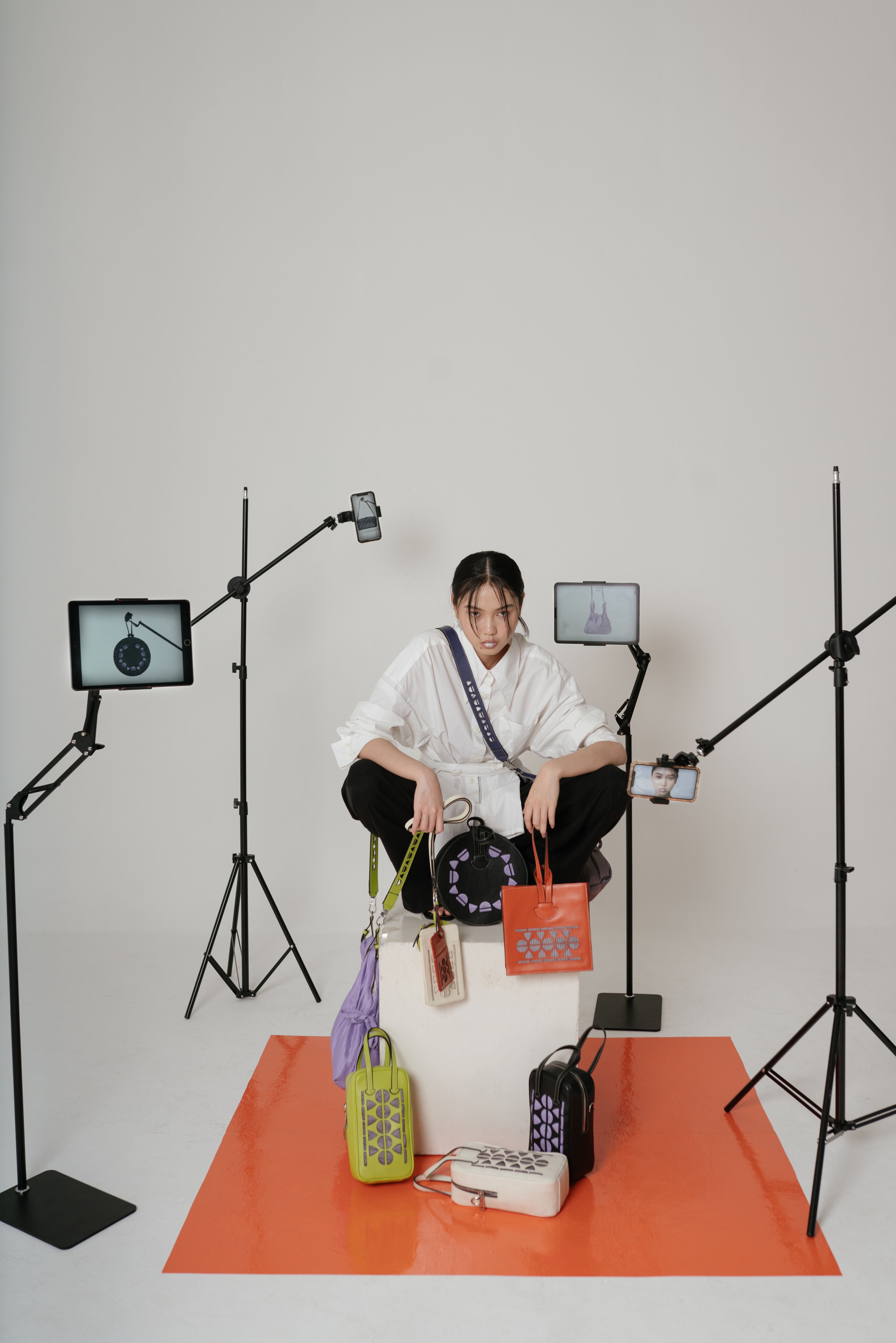 A model sits on an orange square mat, surrounded by camera equipment, posing with a collection of colorful handbags.