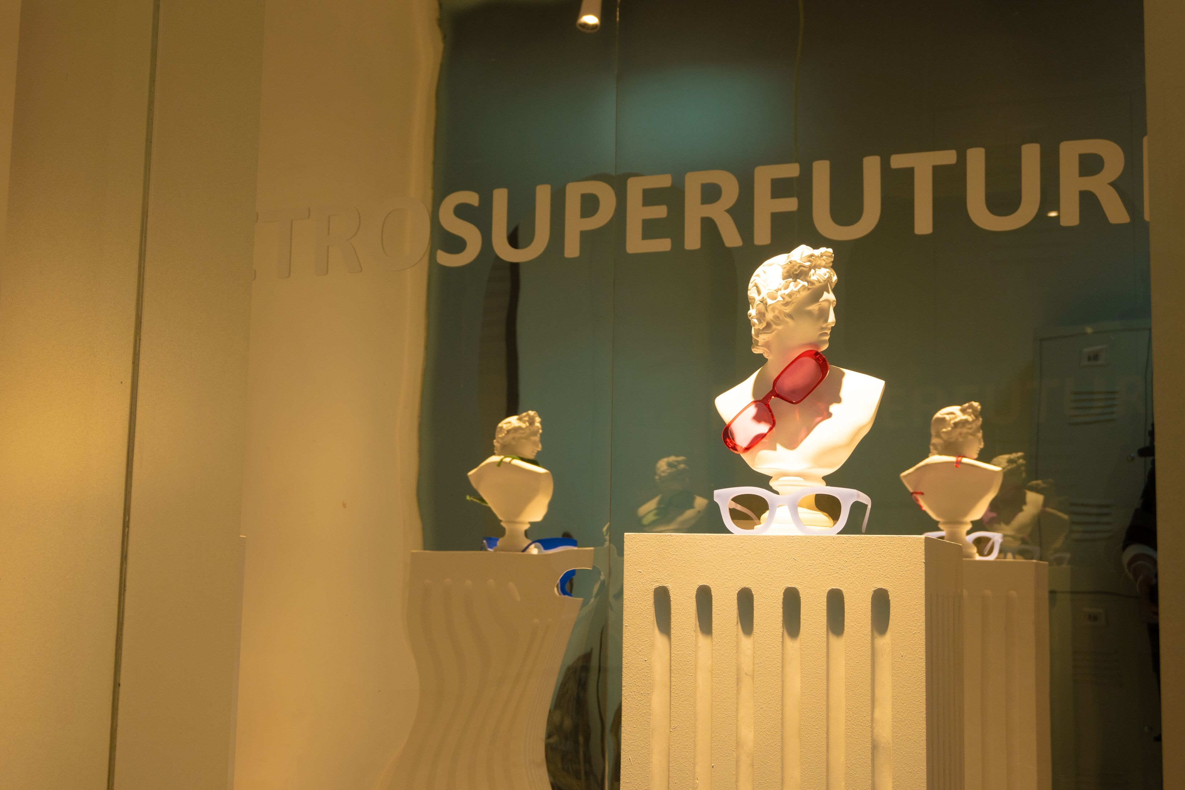 A contemporary boutique window display juxtaposes classical white busts adorned with vibrant modern sunglasses.