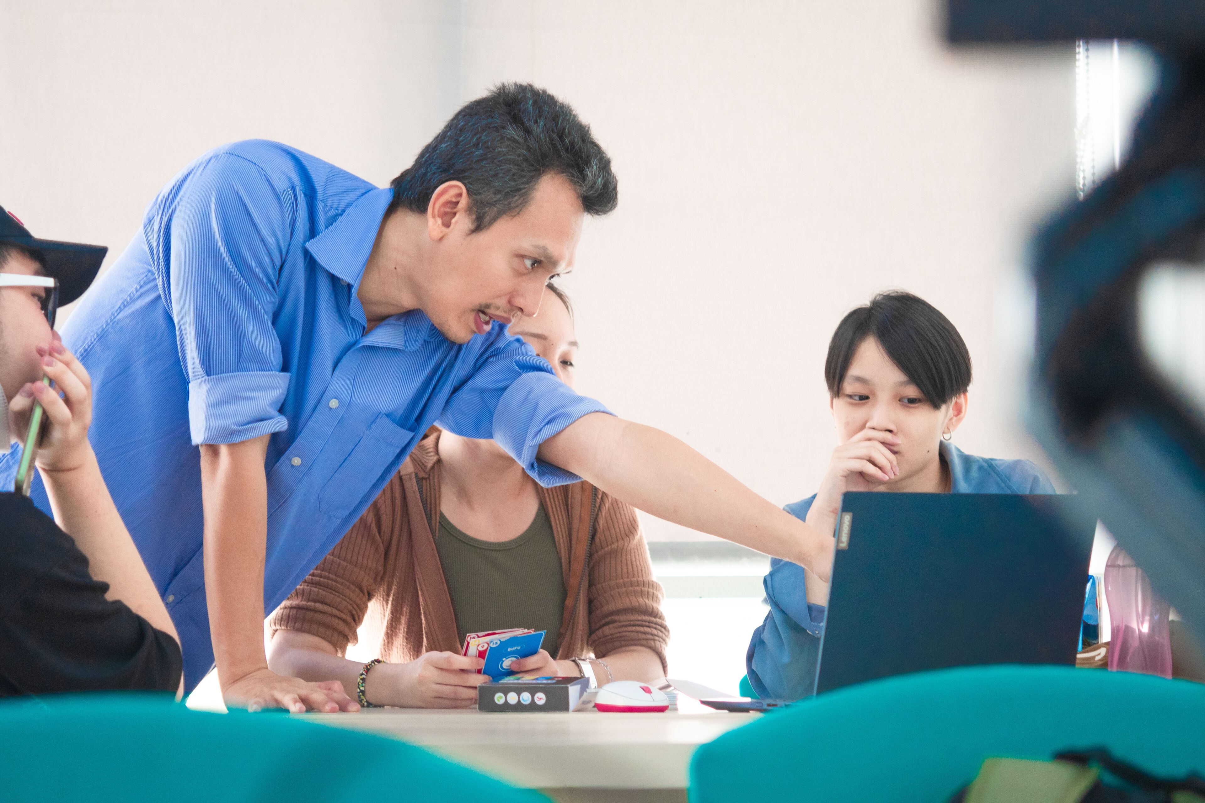 A male teacher in a blue shirt engages with students during a classroom discussion, pointing at a laptop screen while a female student observes intently.