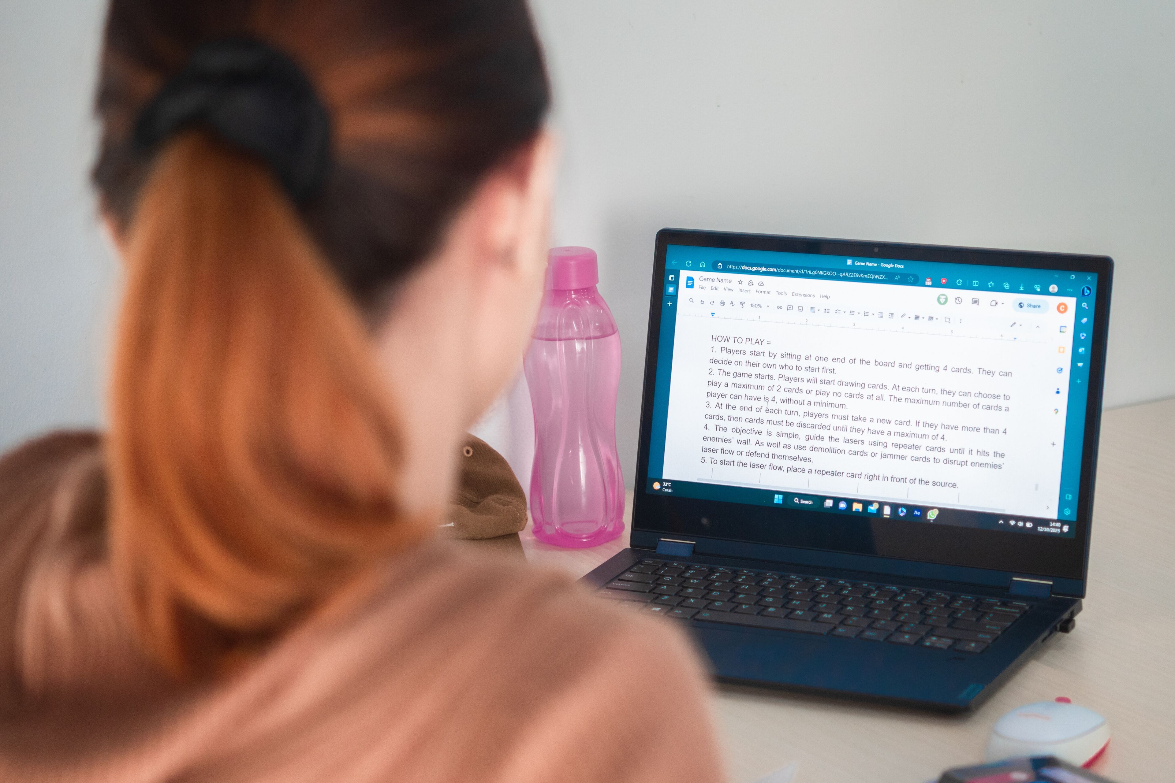 Over-the-shoulder view of a woman with a ponytail using a laptop, displaying a text document about game rules.