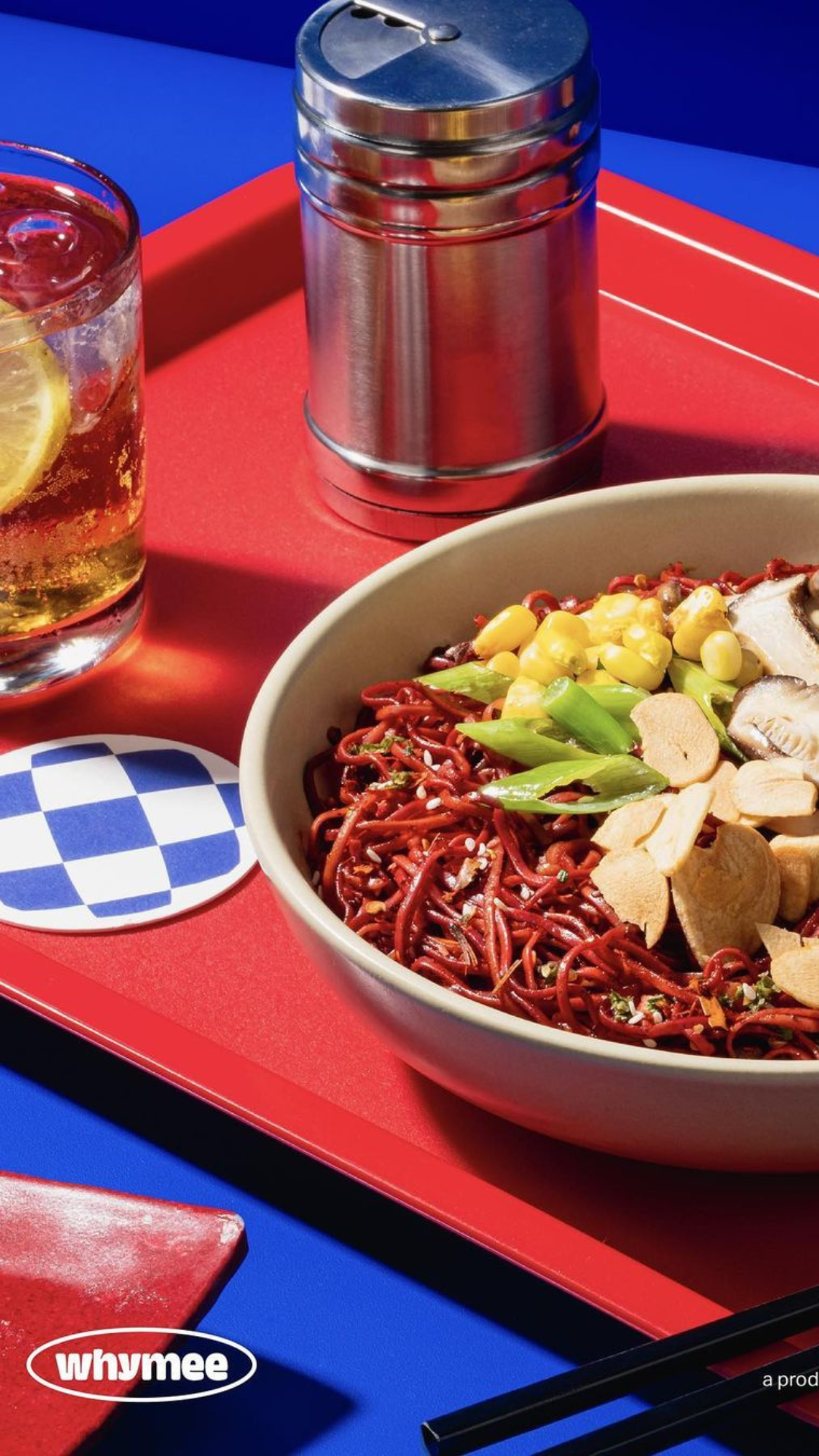An eye-catching food presentation showcasing a bowl of red noodles topped with corn and sliced meats, beside a glass of iced tea on a red tray over a blue table.