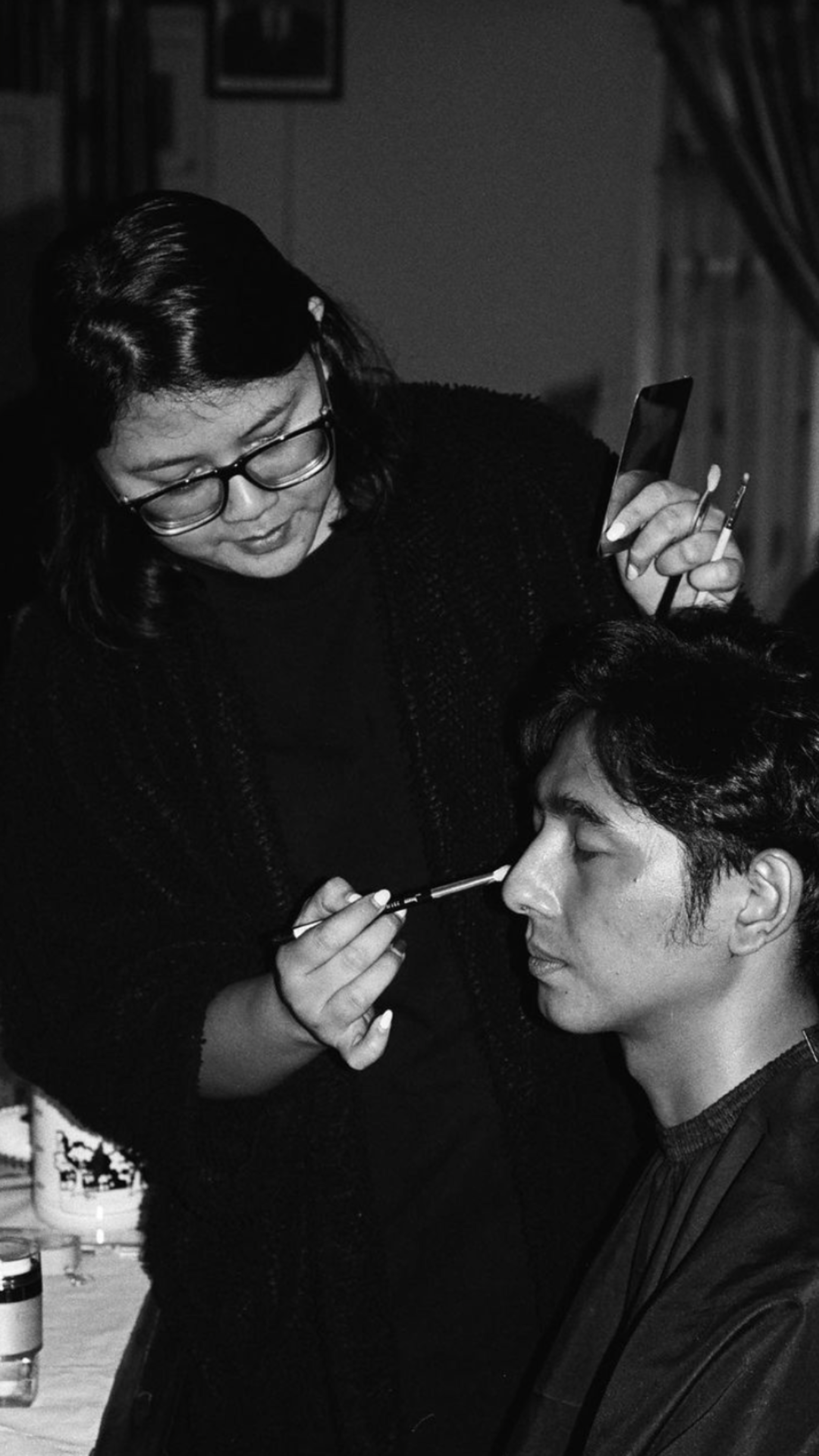 A focused makeup artist applies makeup to a seated man in a black and white setting.