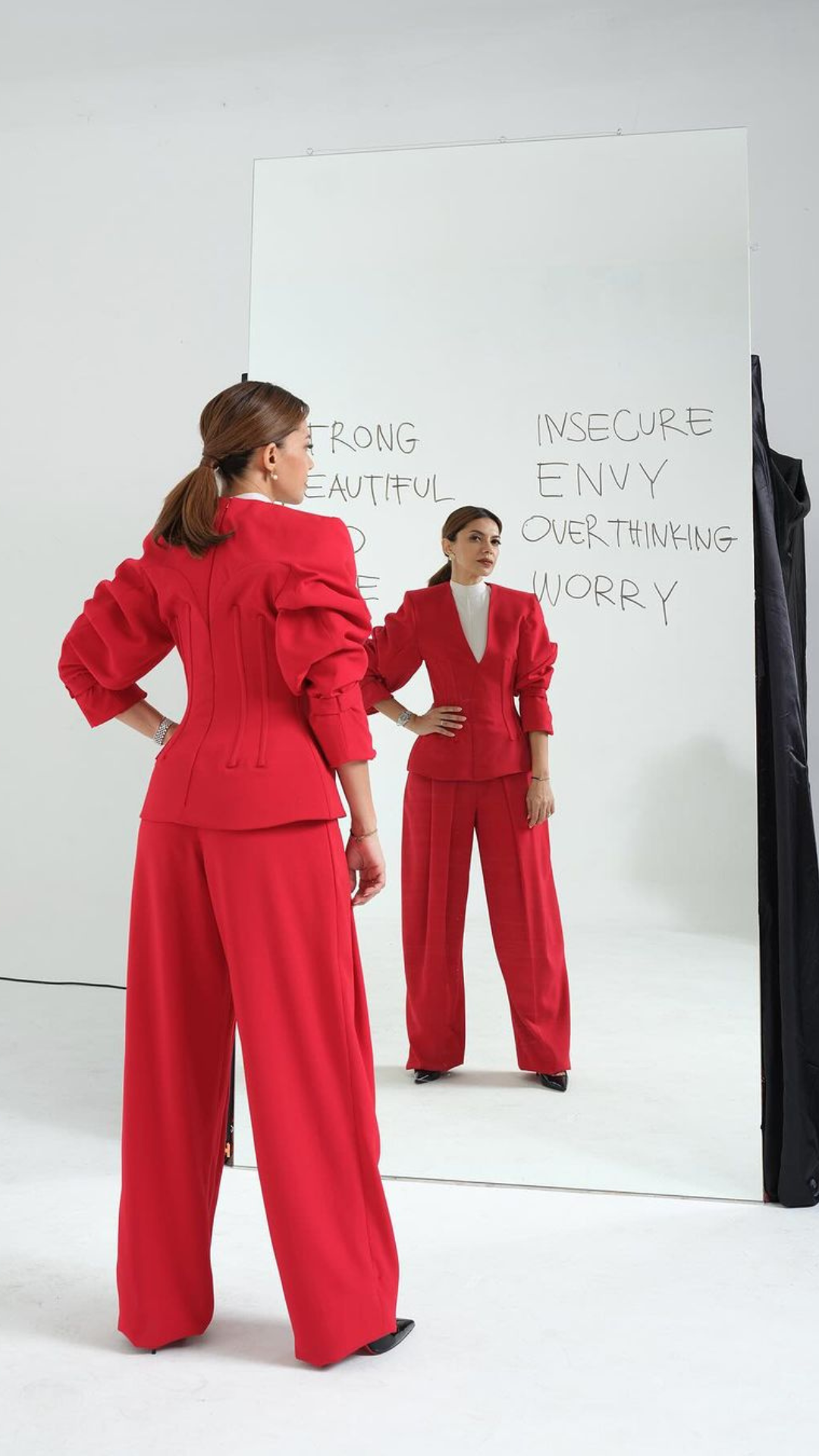 A woman in a red power suit poses before a mirror with words of empowerment and self-reflection written on it.