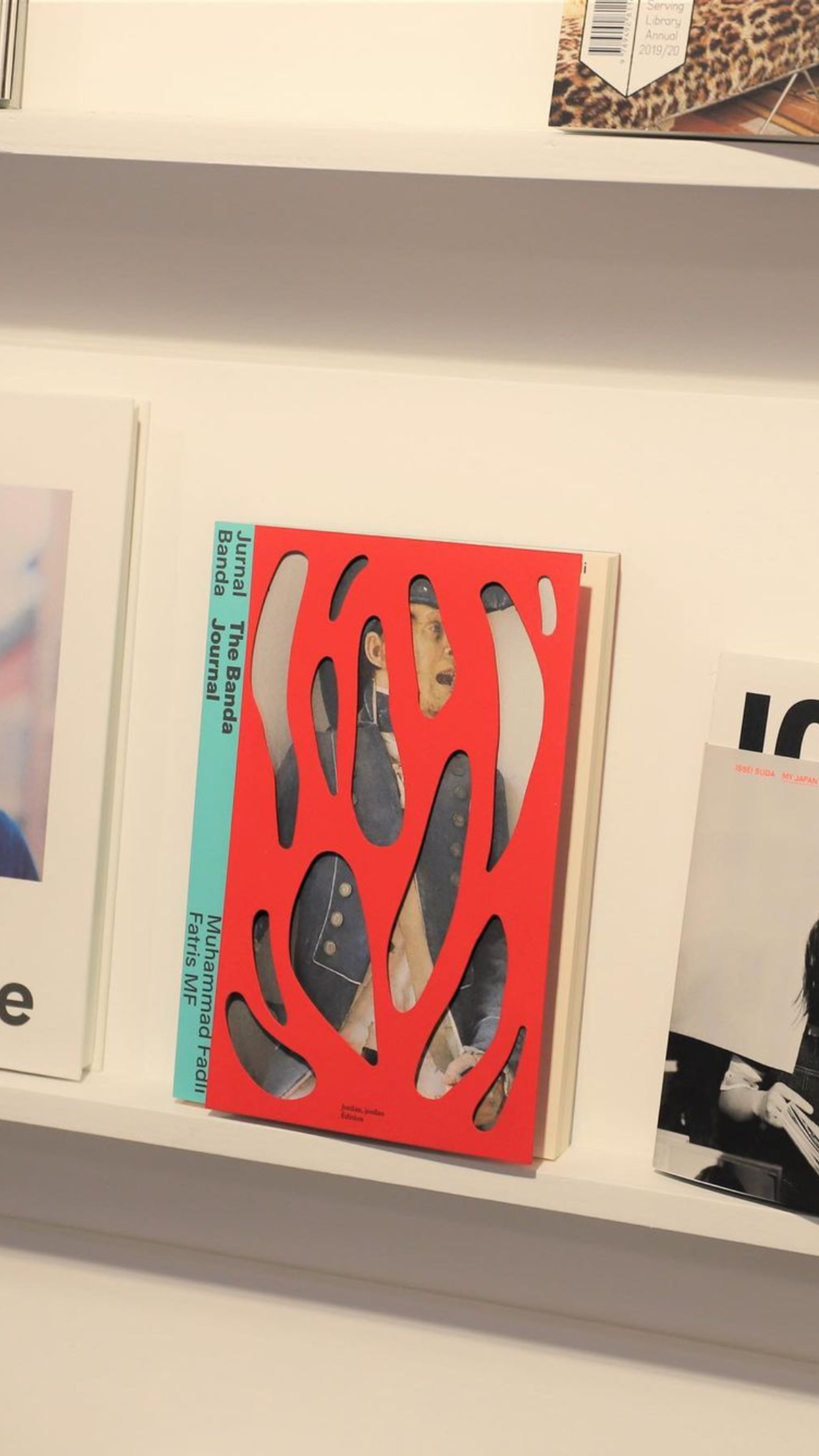 A book with a red abstract cover design featuring cut-out shapes revealing photographs beneath, displayed on a white shelf.