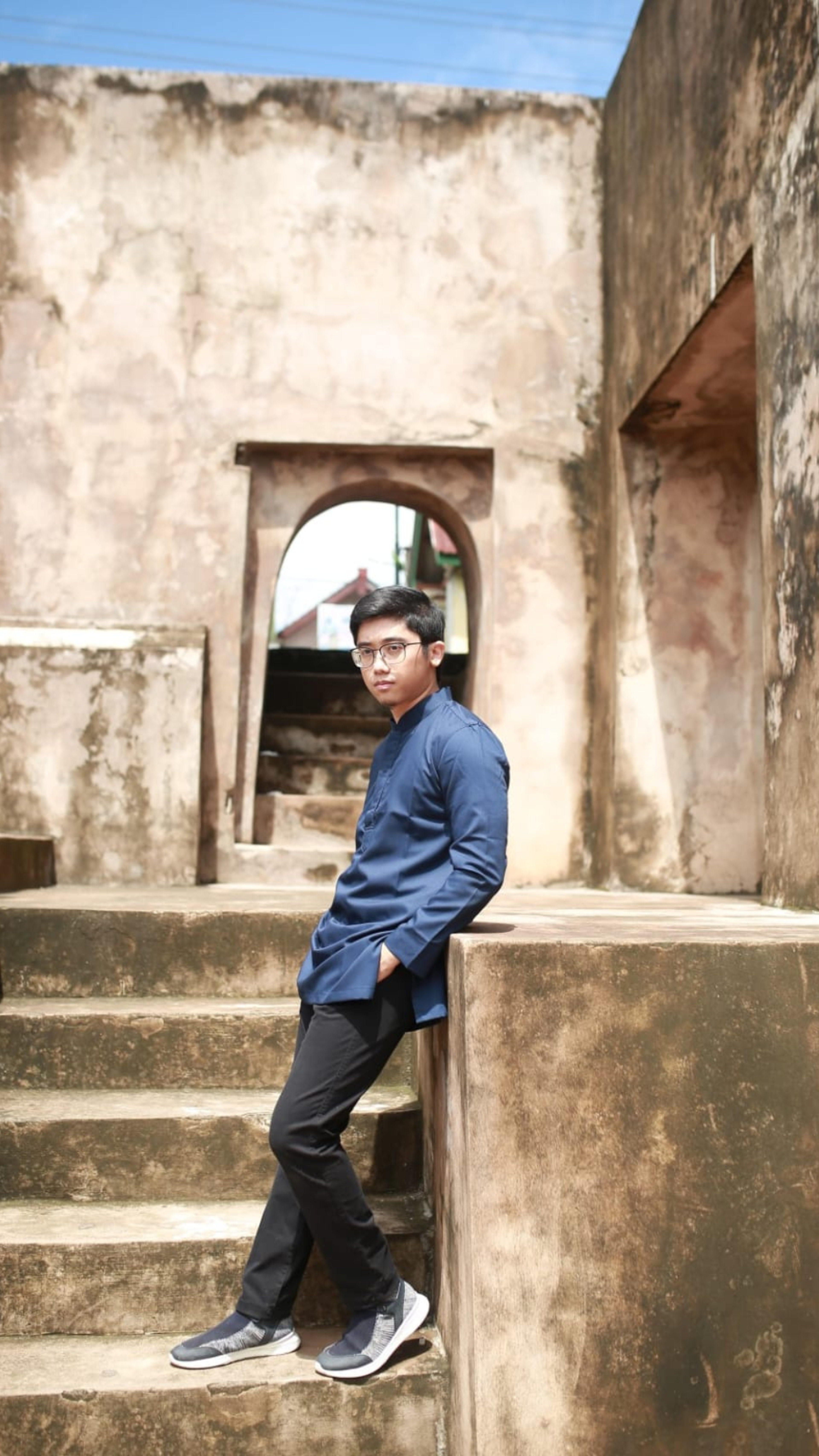 A thoughtful young man in a navy shirt and black pants leans against weathered stone steps of an old building.