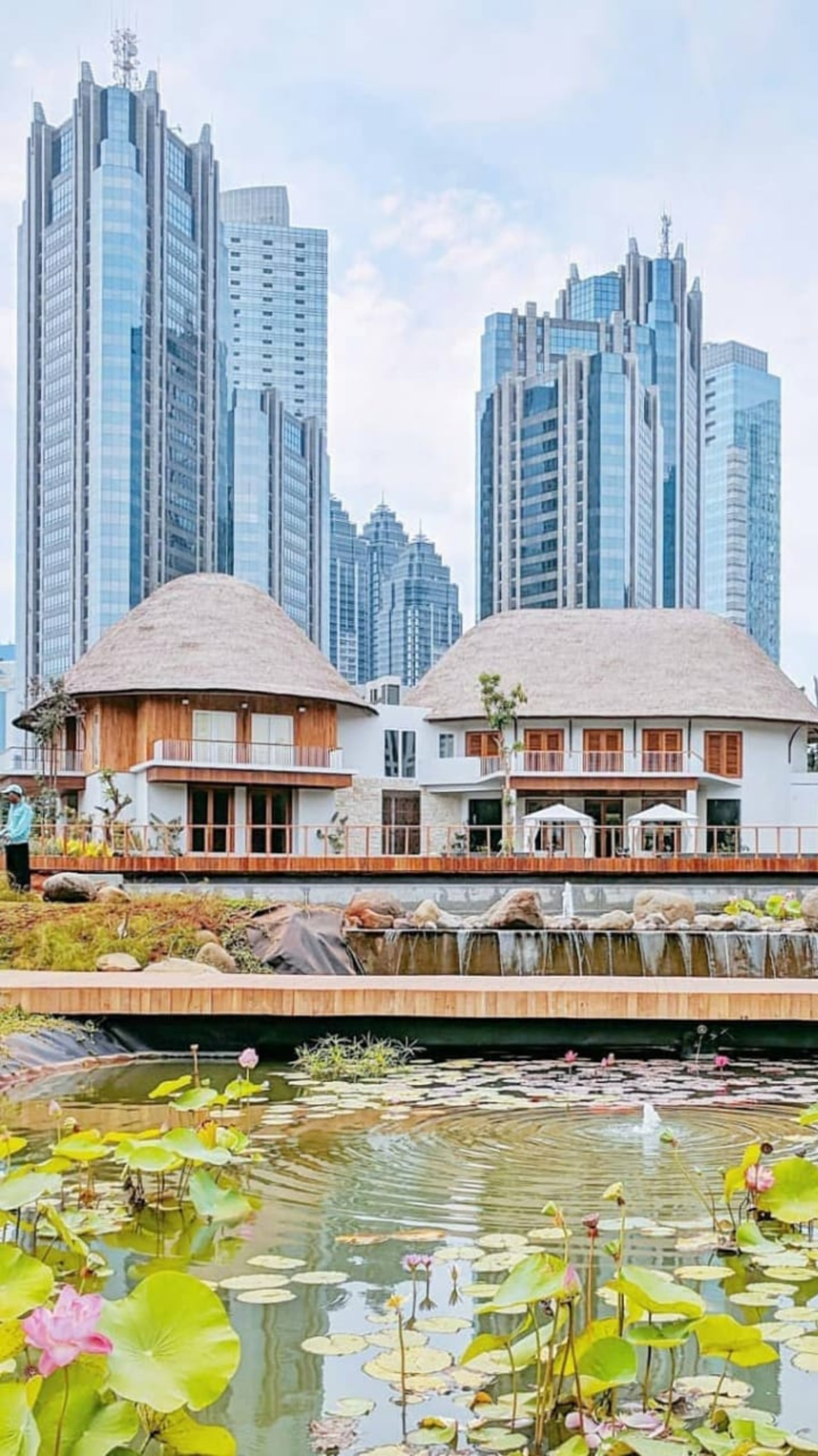 Traditional thatched-roof huts contrast with towering modern skyscrapers, reflecting a blend of old and new architecture by a tranquil city pond with blooming lotuses.