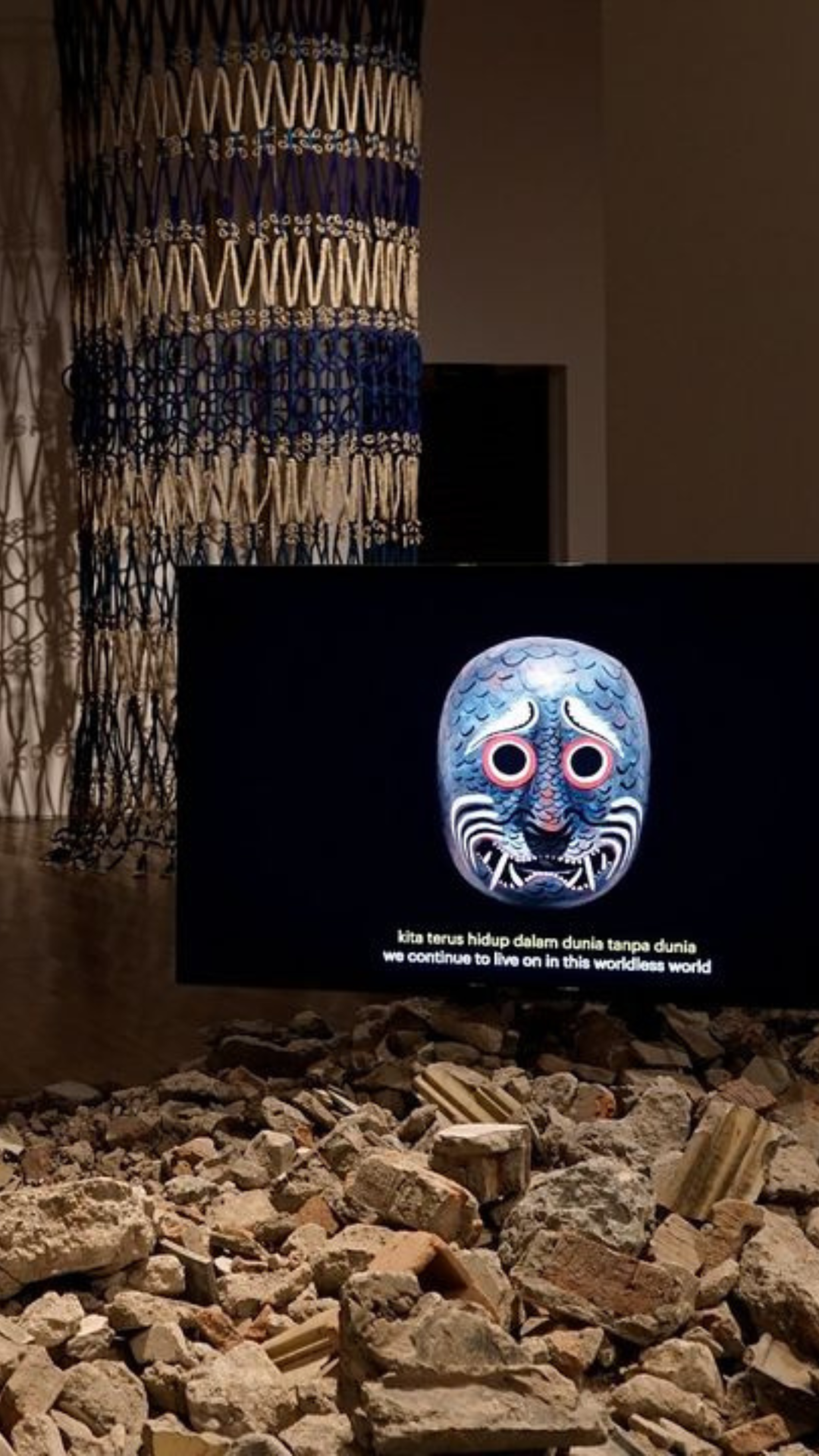 Une installation artistique présentant un masque décoré sur un écran au-dessus des décombres, avec une phrase en indonésien et sa traduction anglaise affichée.