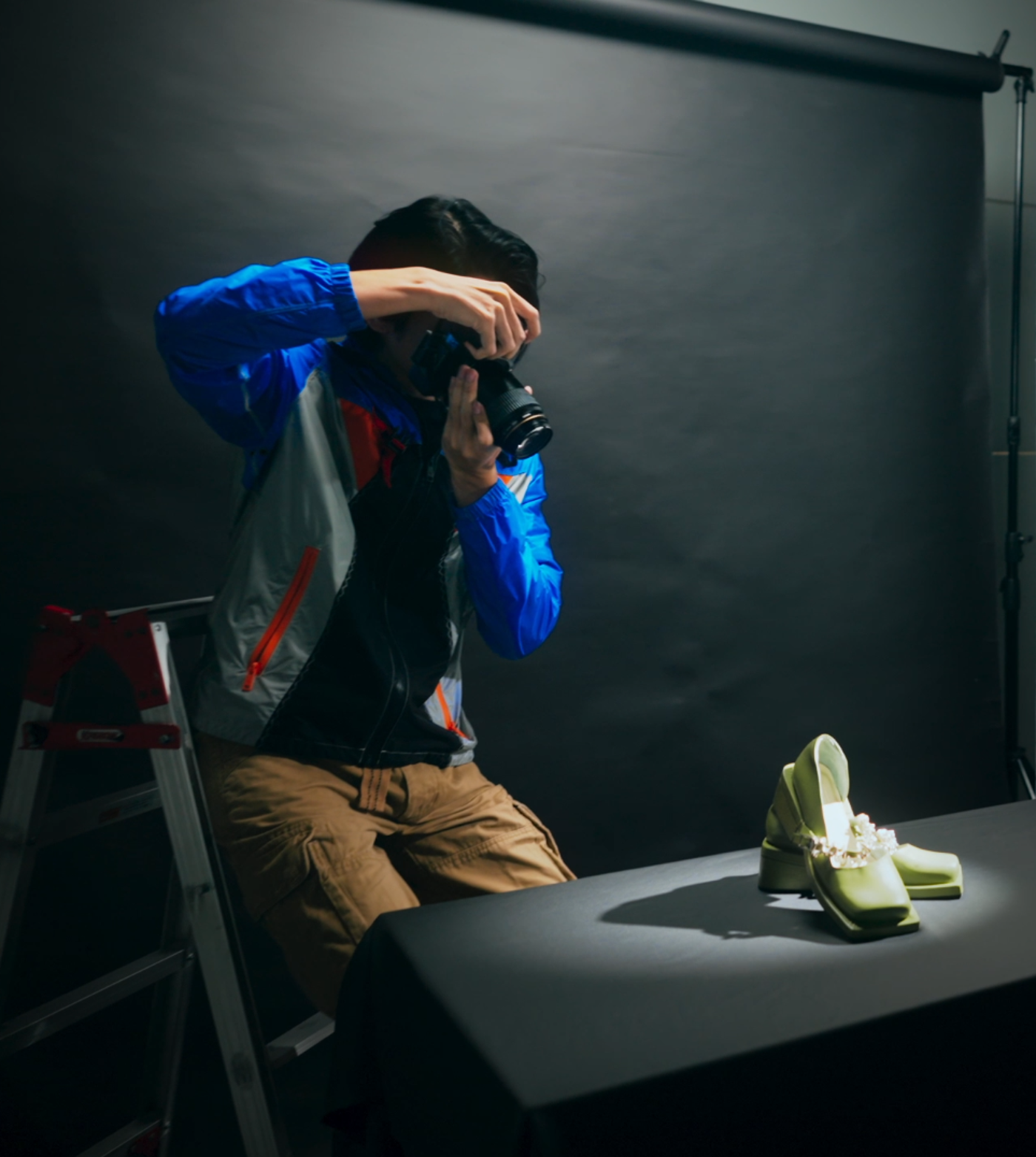 A photographer is taking a picture of a pair of green shoes in a studio setting. The photographer is using a camera and is positioned on a ladder.