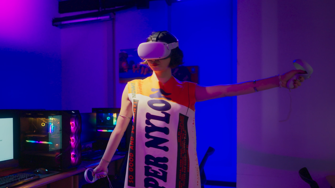 A student wearing a bold graphic dress uses a VR headset and controllers in a dark, neon-lit room with high-performance PCs. The setting showcases innovative digital interaction at LCI Jakarta, blending fashion and technology.
